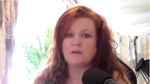 A redhaired woman speaks into a microphone. She's slightly backlit from the sun behind her and a fern is visible over one shoulder.