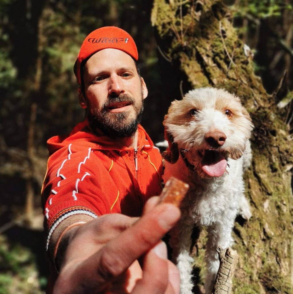 Dog behaviour - Photo of a bearded man wearing red hat and jumper holding a small white dog, stood in front of a tree.