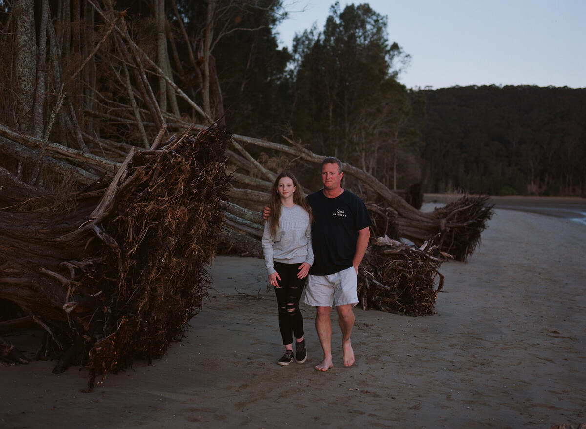 Raeden, 13, walks with her father Jim, 47, next to driftwood washed up on a beach in Batemans Bay where they now live. Image: Matthew Abbott / Save the Children