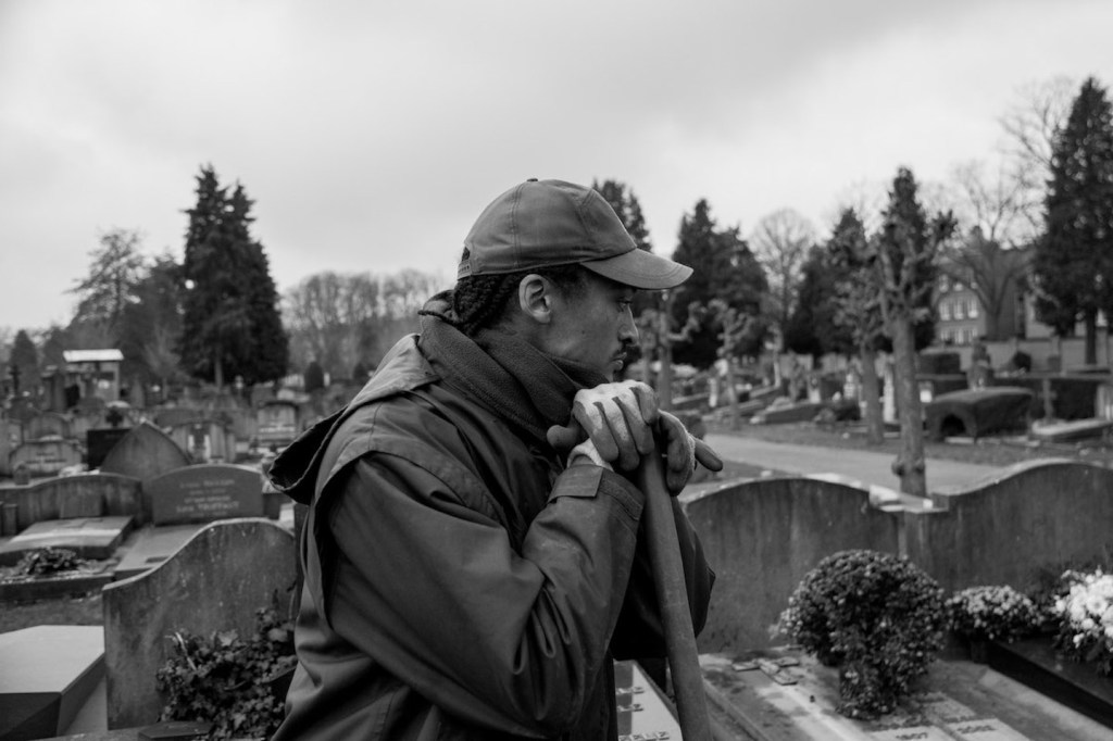 Jacques Vermeer, Patrick, cemetery - Black and white photo of a man in a cap resting his head on a spade in a graveyard.