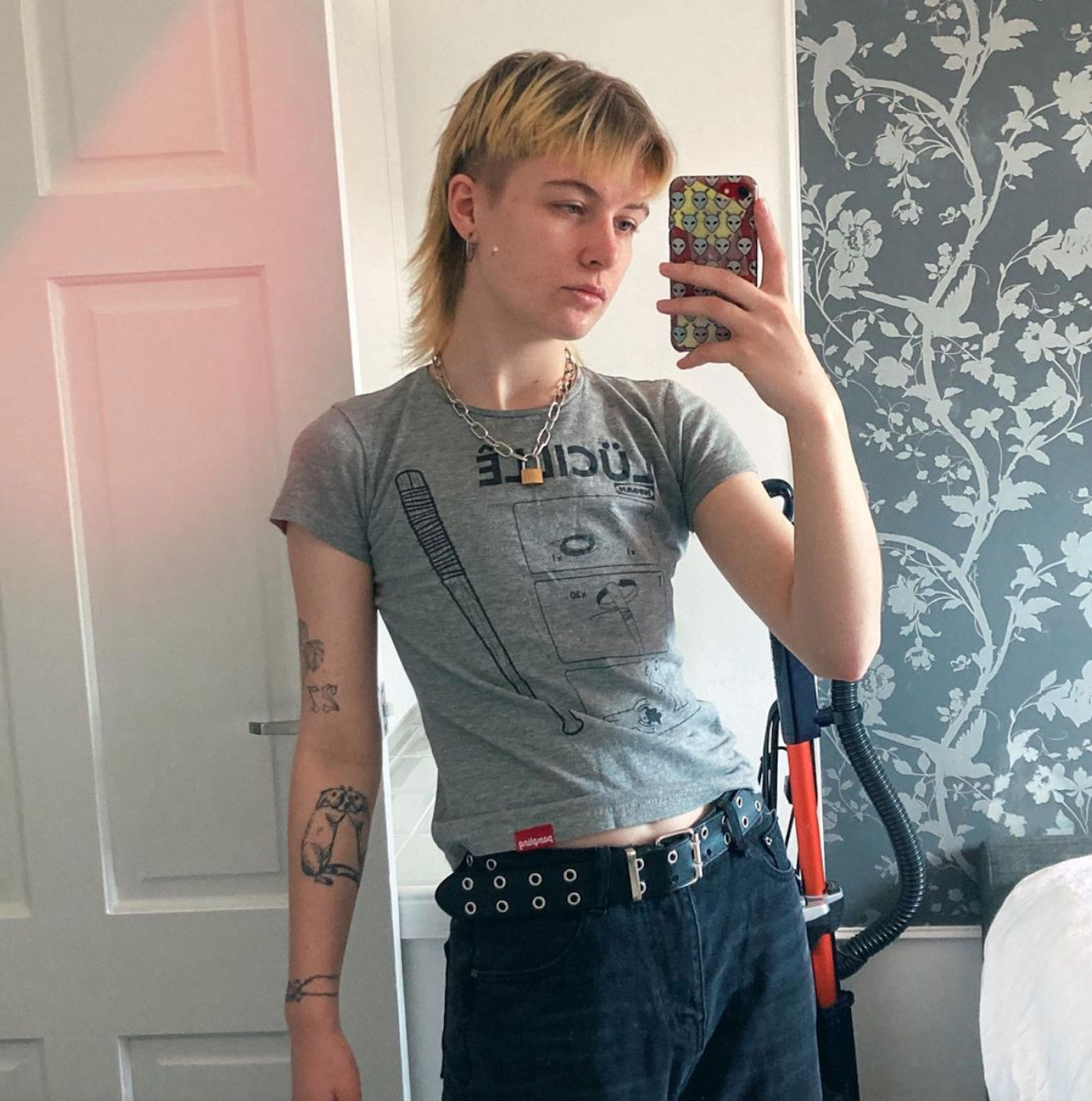 Eine junge Person mit blondem Vokuhila, dicker Kette, diversen Tattoos und grauem T-Shirt macht in einem Spiegel ein Selfie