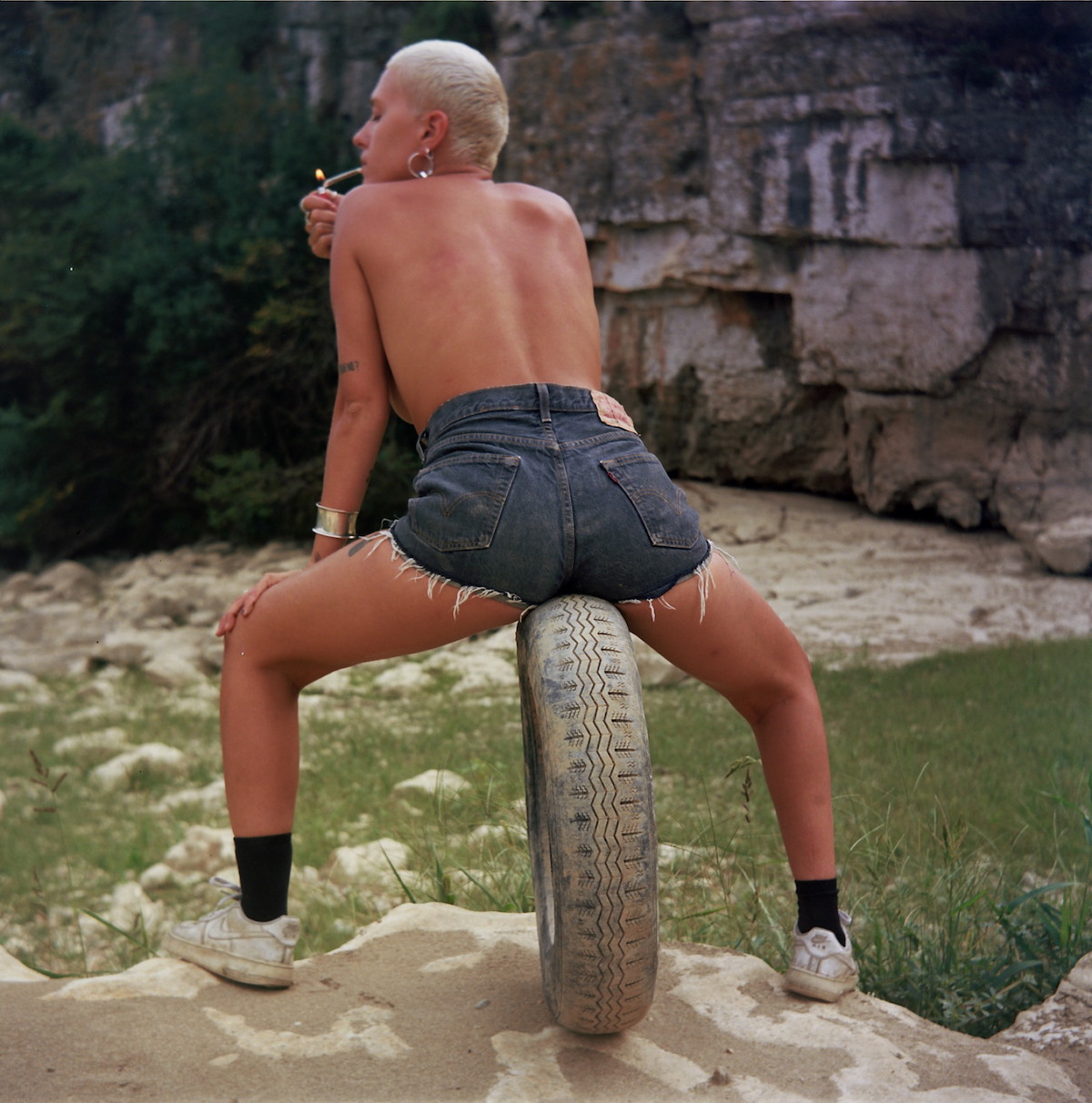 Of Manifesto, Belgium, porn - A blonde figure in denim hot pants sits on a tyre, smoking a cigarette.