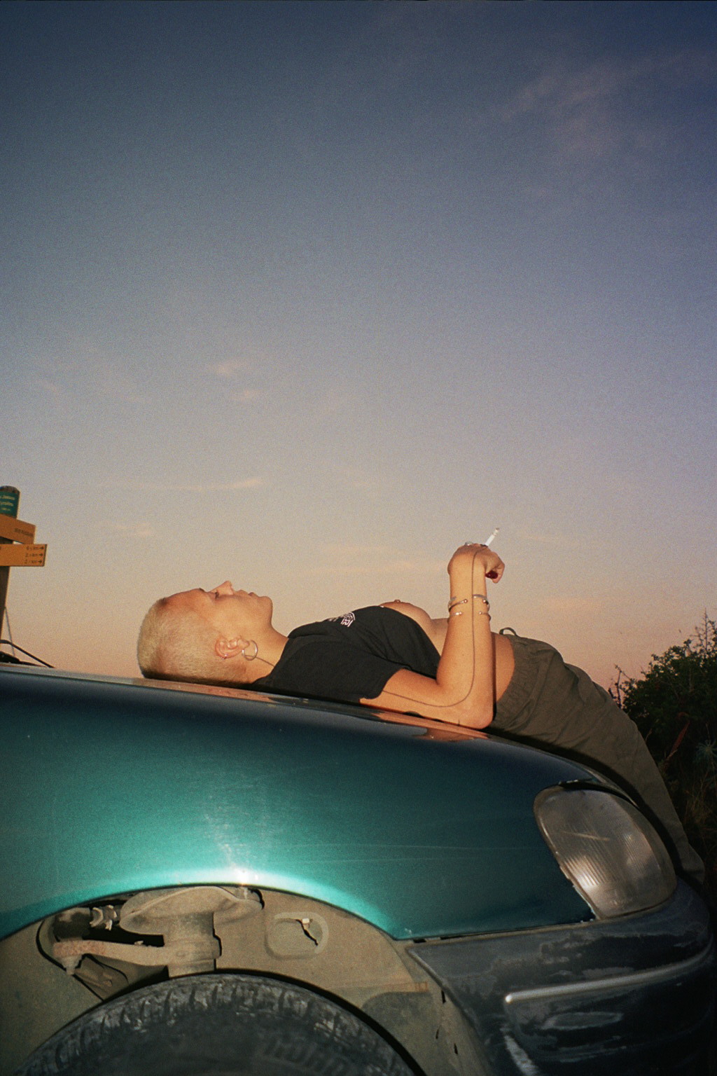 Of Manifesto, Belgium, porn - Photograph of blonde person lying on the hood of a green car at sunset smoking a cigarette.