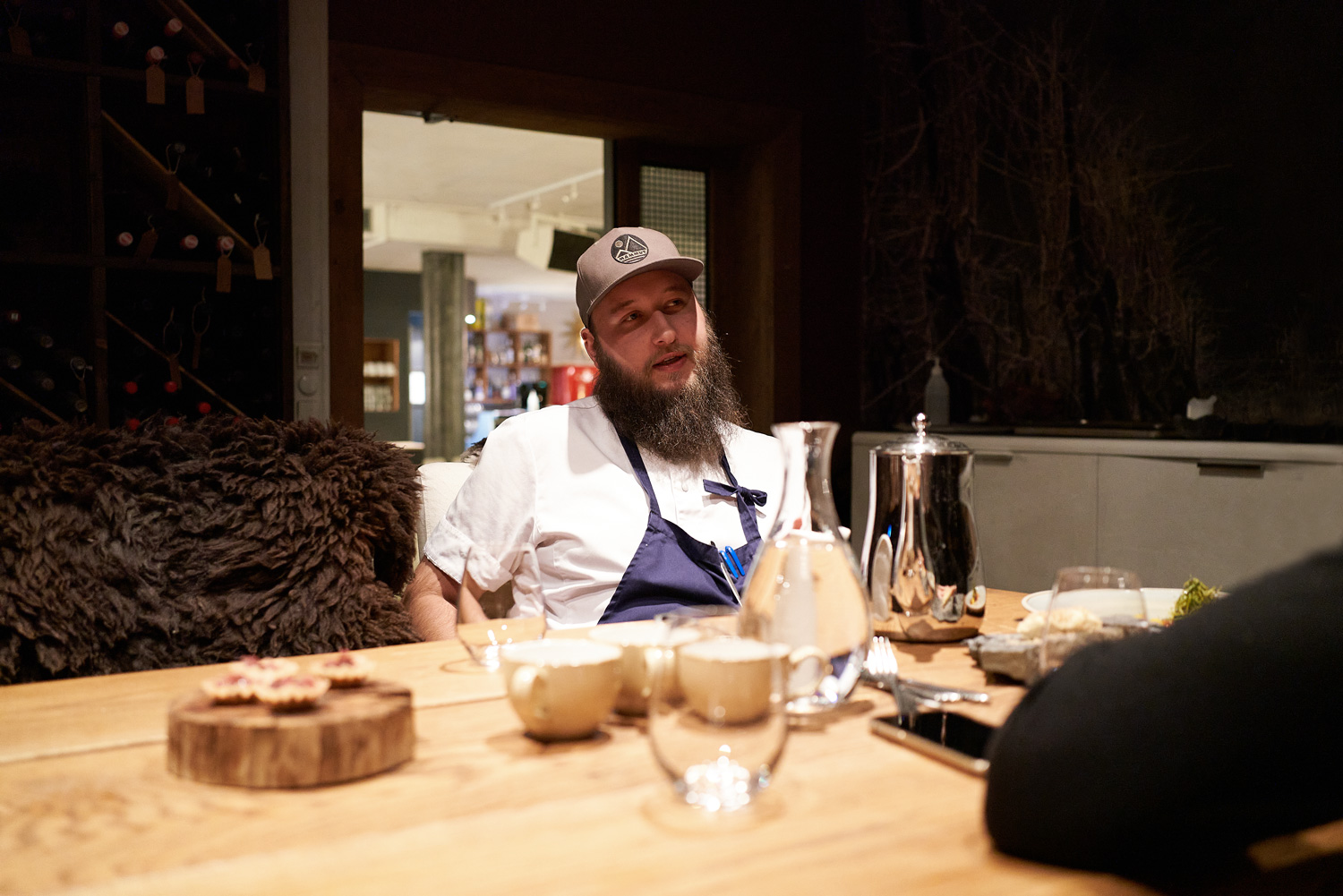 Arctic cuisine, Svalbard, Nick Casini - Photo of a bearded man in a baseball cap and a chef's apron sat at a wooden table.