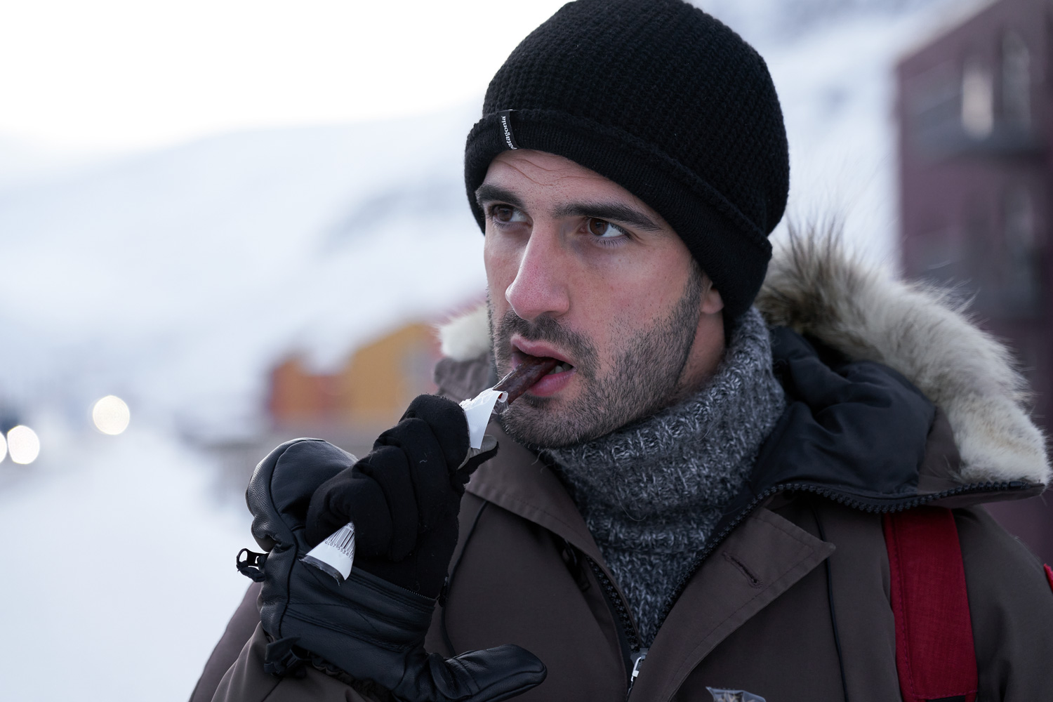 Arctic cuisine, Svalbard, Nick Casini - Photo of man wearing black beanie hat, and a brown parka coat eating a brown stick of meat.