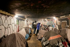 Paris refugees Porte Brunet – tunnel covered in graffiti with two rows of tents and a muddy path in the middle. At the back, a group of young kids is standing around.