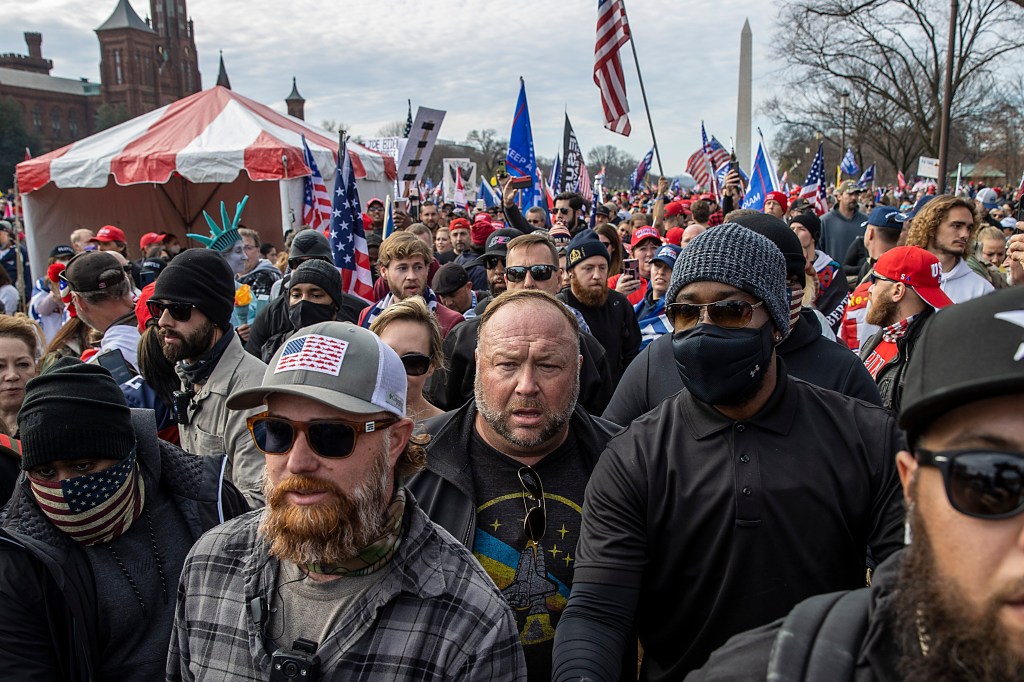 Alex Jones is seen in a crowd of people, wearing a gray t-shirt with a rocket ship on it and looking befuddled.