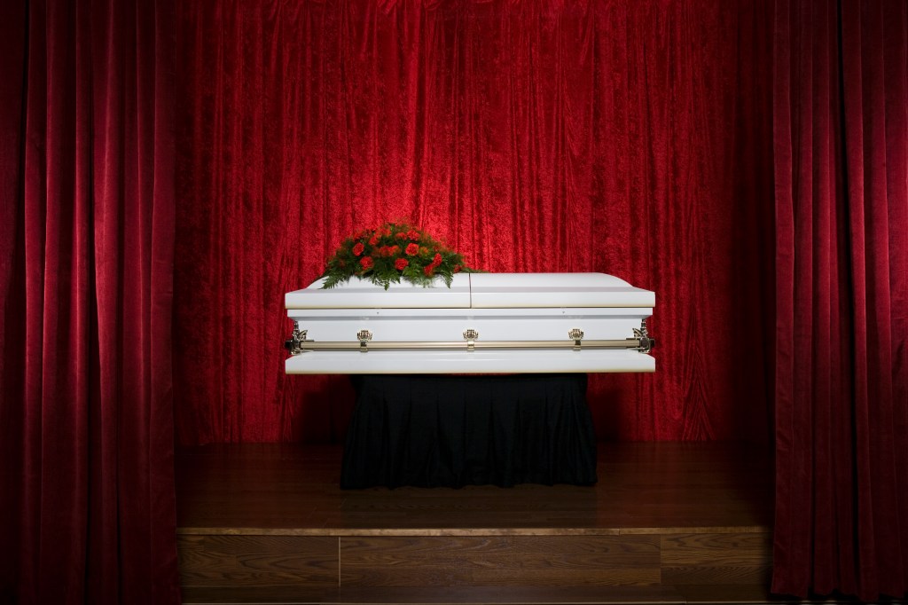 A white casket sits on a wooden stage, backed by red velvet curtains. A bouquet of red roses sits on top of the casket.