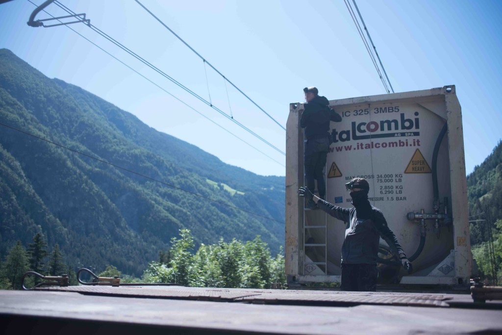 GifGas, trains, Belgium - Photo of a man stood on the back of a freight train carriage pointing at a mountain uncder a blue sky.