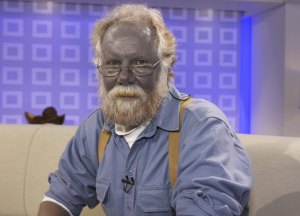 A blue-tinged man in suspenders and glasses and a blue workshort gazes dolefully at the camera. He's sitting on a white couch.