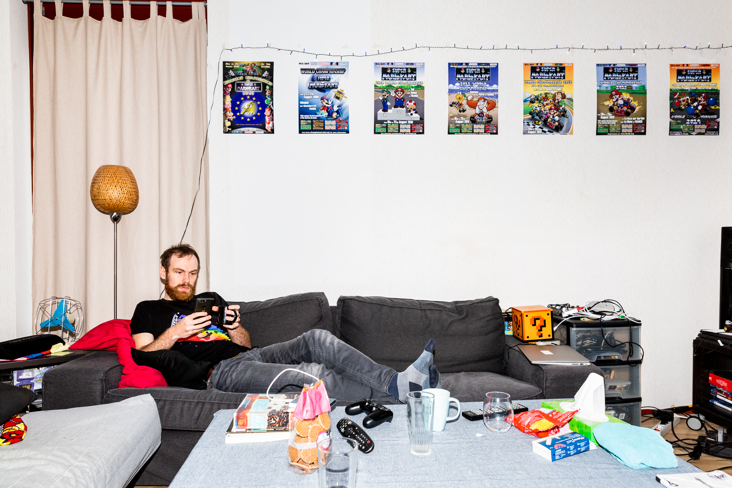 Speedrunning, Marieka de Bra, gaming - Photograph of a man laying on a black sofa looking at his phone, posters for Mario Kart championships above him.