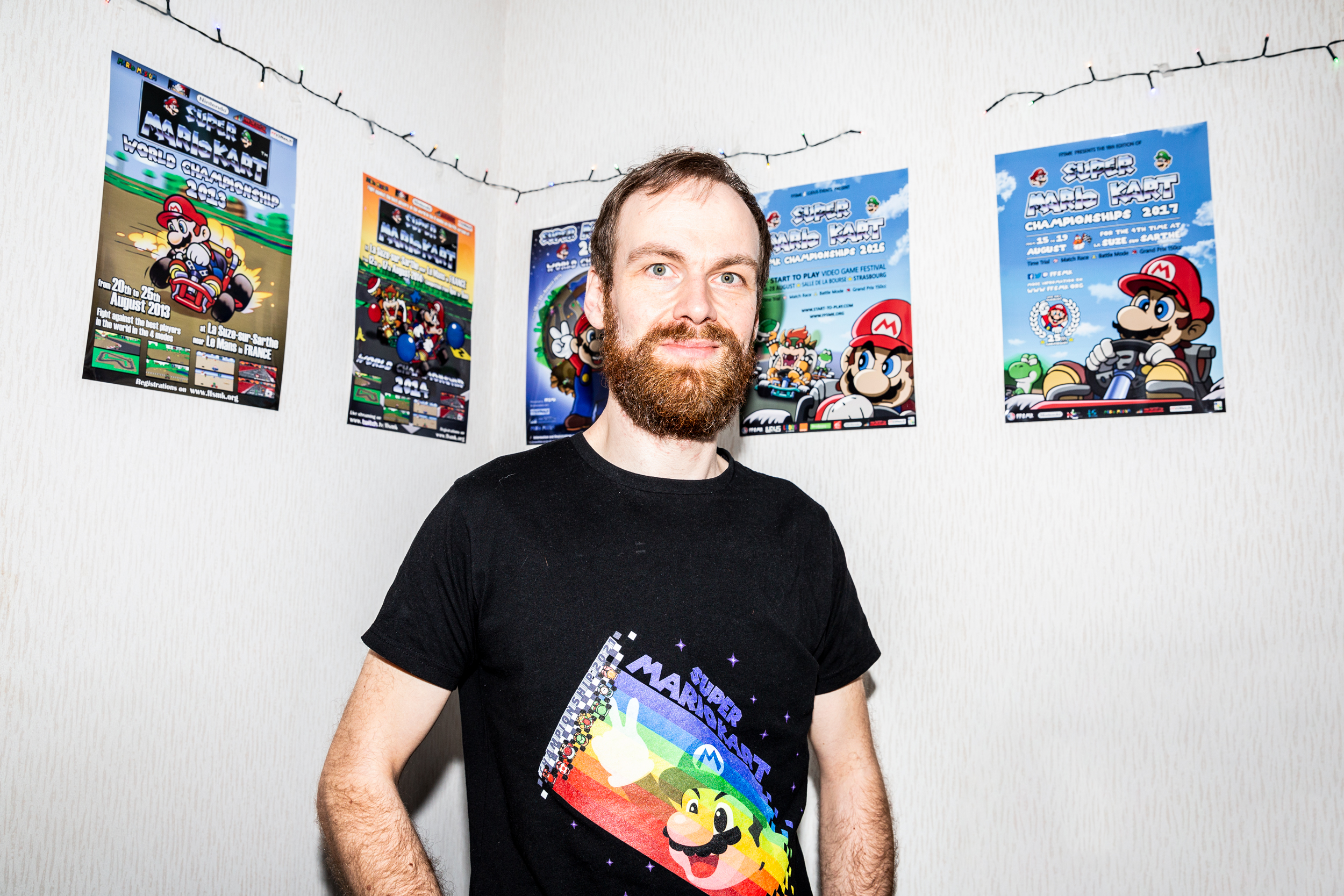 Speedrunning, Marieka de Bra, gaming - Photograph of a bearded man in a black t-shirt stood under a collection of Mario Kart posters.