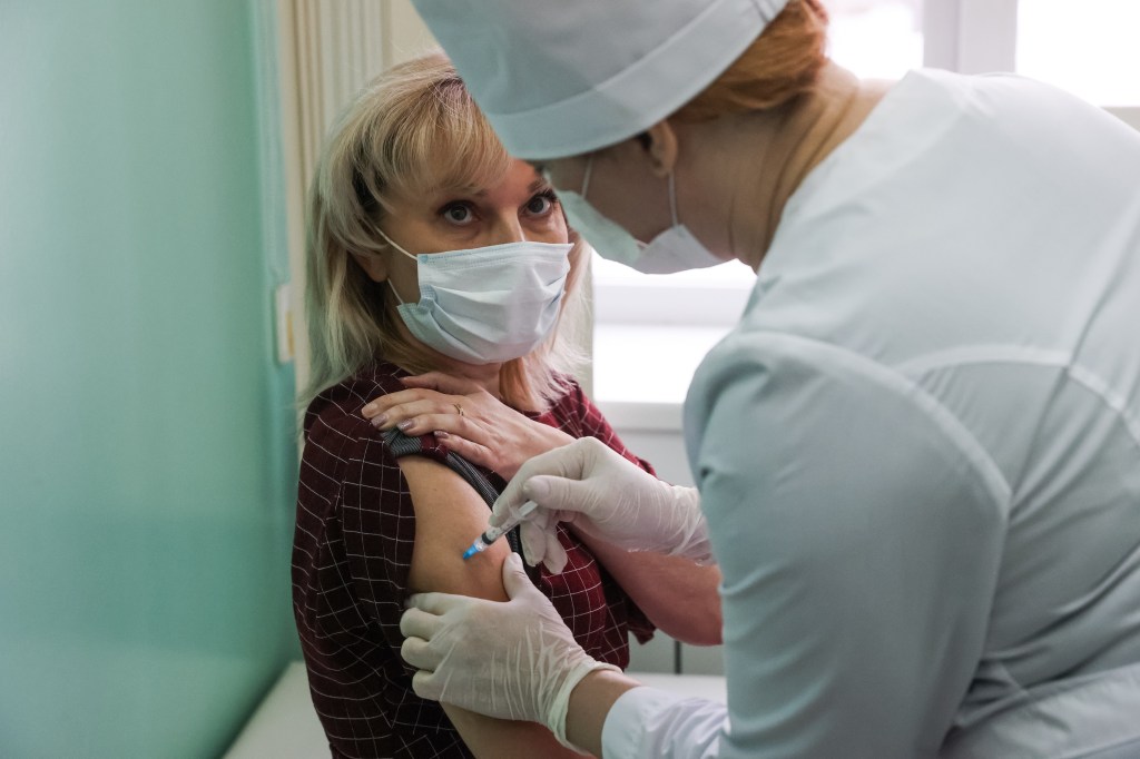 A blonde woman in a white mask receives a shot in her arm from a nurse in a white hat.