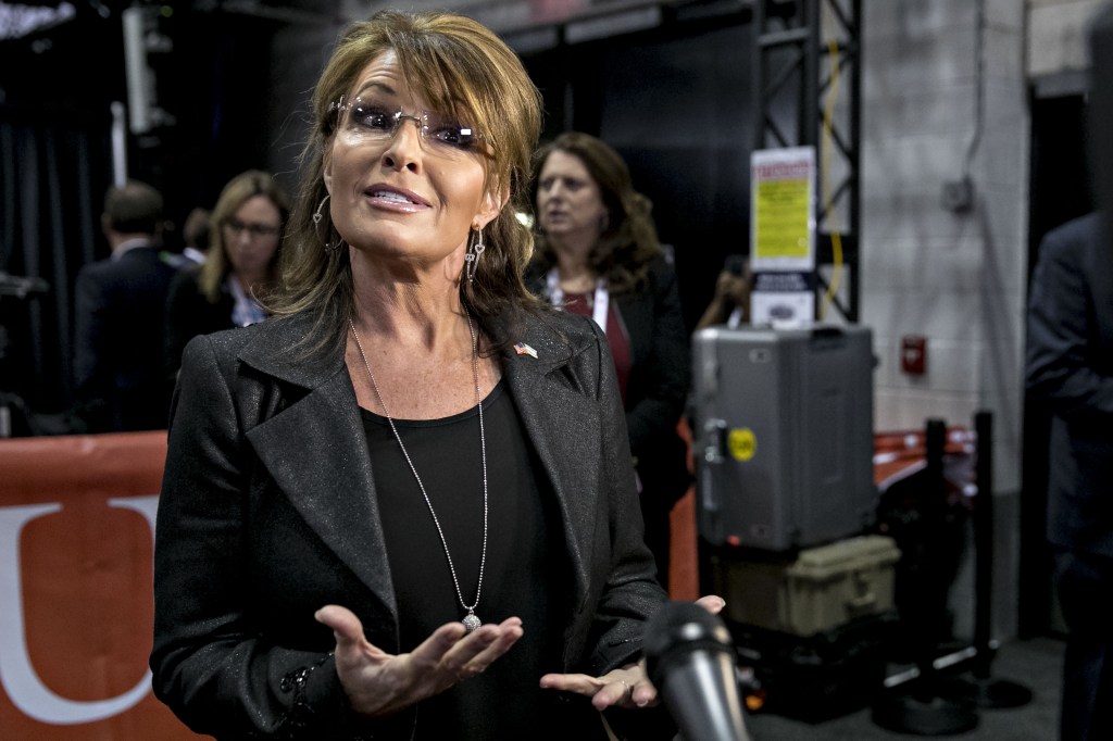 Sarah Palin, wearing glasses, a black shirt, a blackblazer and a necklace, speaks towards the camera, gesturing with her hands.