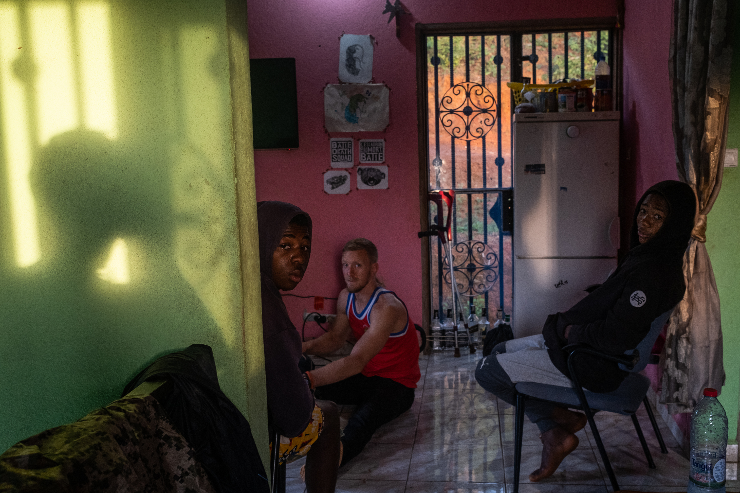 Foundation Ngannou, Batié, Cameroon – Two kids sitting on chairs while sam is sitting on the floor and doing something. They're in the kitchen, a room with pink walls next to another room with light green walls.