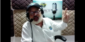 Image shows a man in a white shirt, black baseball cap, black beard and black glasses speaking into a microphone. He has headphones on and is raising one fi