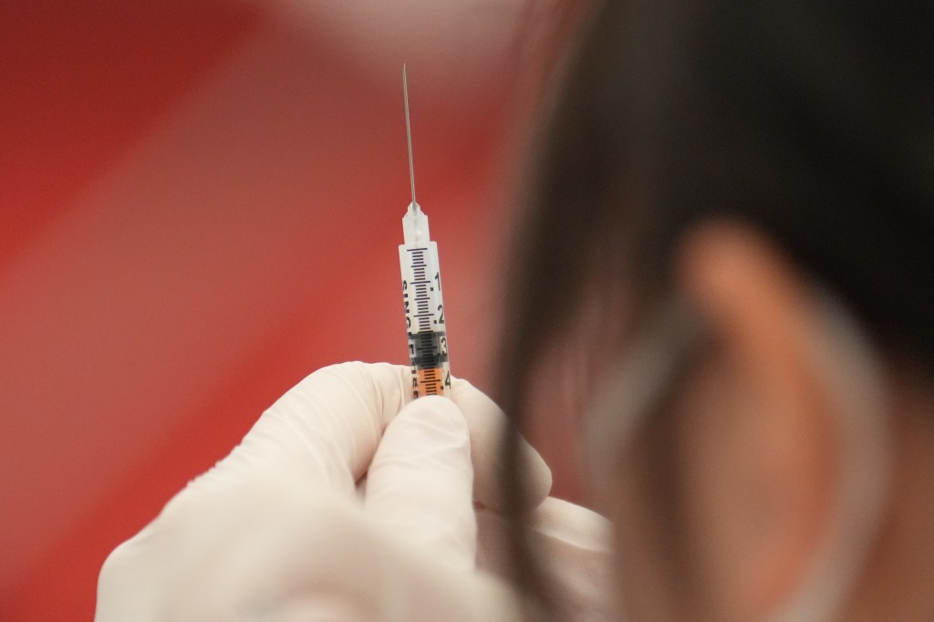 Photo shows a white gloved hand holding a syringe - a person's head with dark hair is also visible near the needle. there's a red background behind the needle.