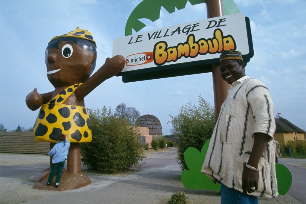 Bamboula's Village – Picture of the entrance of Bamboula's Village. Left: the comic book character, right: a sign reading “le Village de Bamboula”, attached to a cartoon palm. In the foreground, two Black men, one smiling at camera.