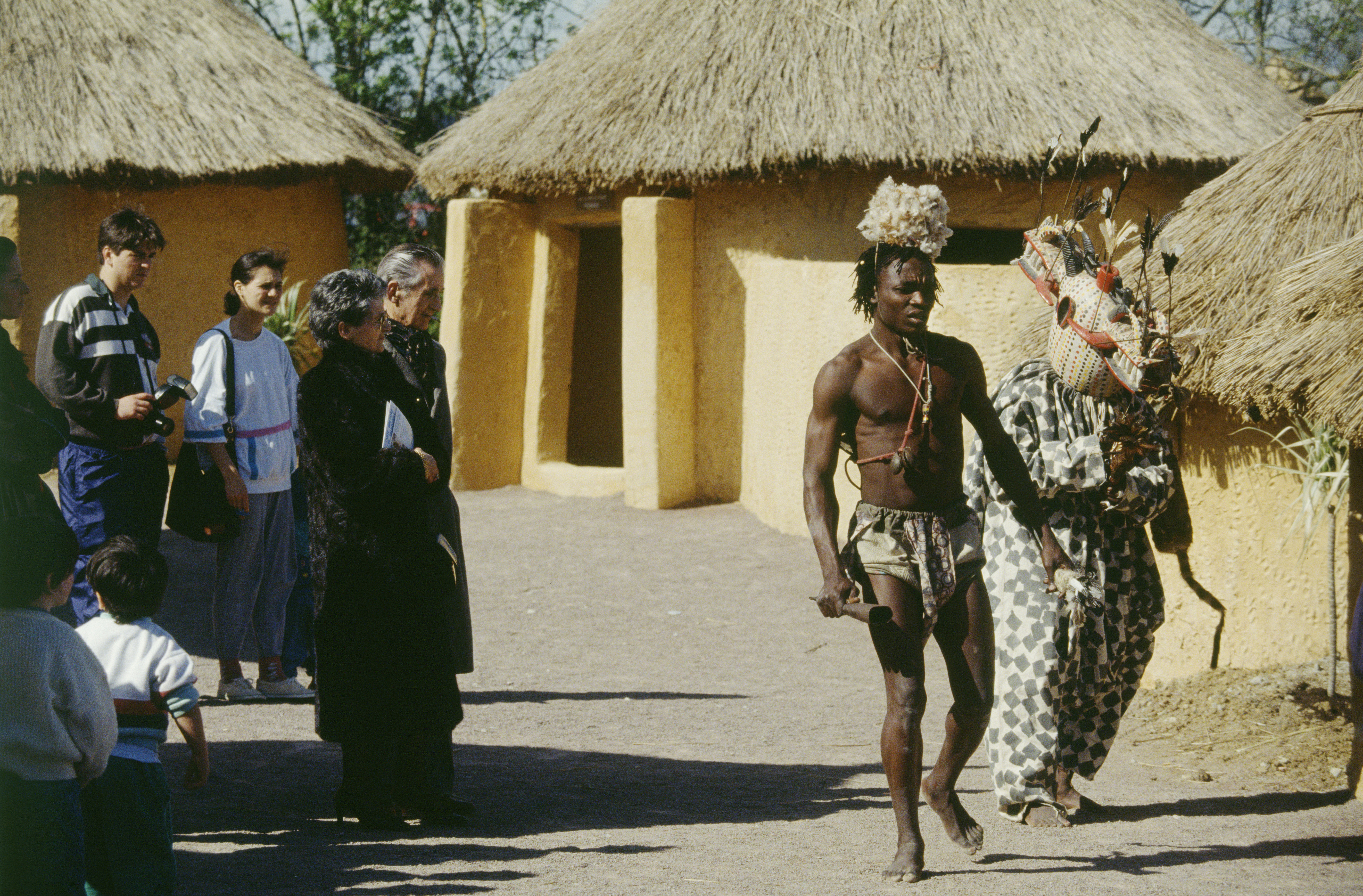 Bamboula's Village – Barefoot man wearing sorts, some necklaces and a straw hat, walking next to someone wearing a ritualistic mask and a printed onesie as the audience watches by.