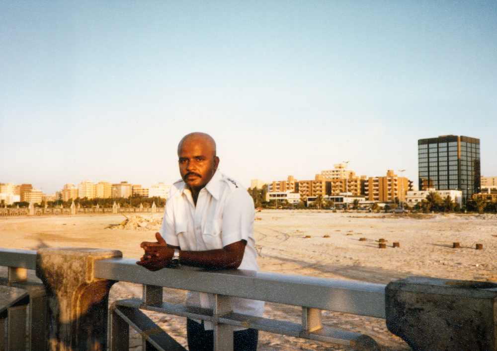 Saleh al-Tamimi, Abu Dhabi – man with a shaved head and a 70s moustache, wearing a white linen shirt and posing in front of an empty lot with low and mid-rise buildings in the background.