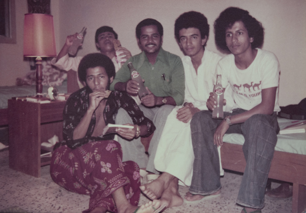 Saleh al-Tamimi, Abu Dhabi – group of men sporting 70s hairstyles sitting in a small room with two single beds and drinking soda.