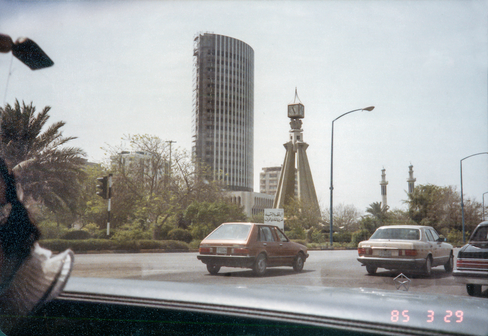 Saleh al-Tamimi, Abu Dhabi – photo taken from inside the windshield of a car, picturing a road near a tall glass building and a clock tower surrounded by greenery.