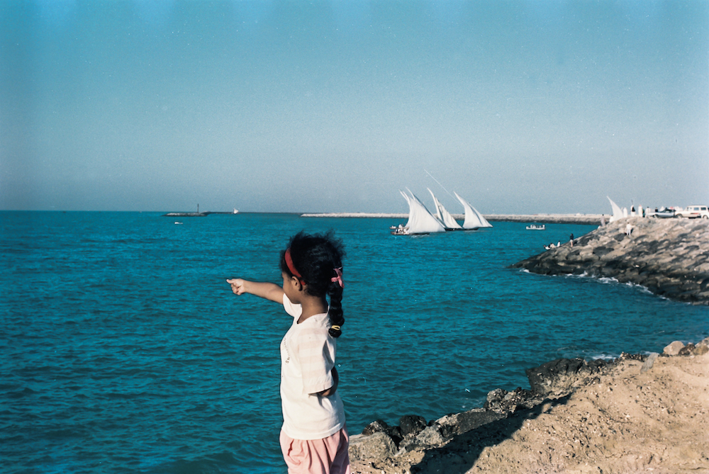 Saleh al-Tamimi, Abu Dhabi – A young child standing on rocks at the seaside, pointing towards the horizon where multiple sail boats are sailing.