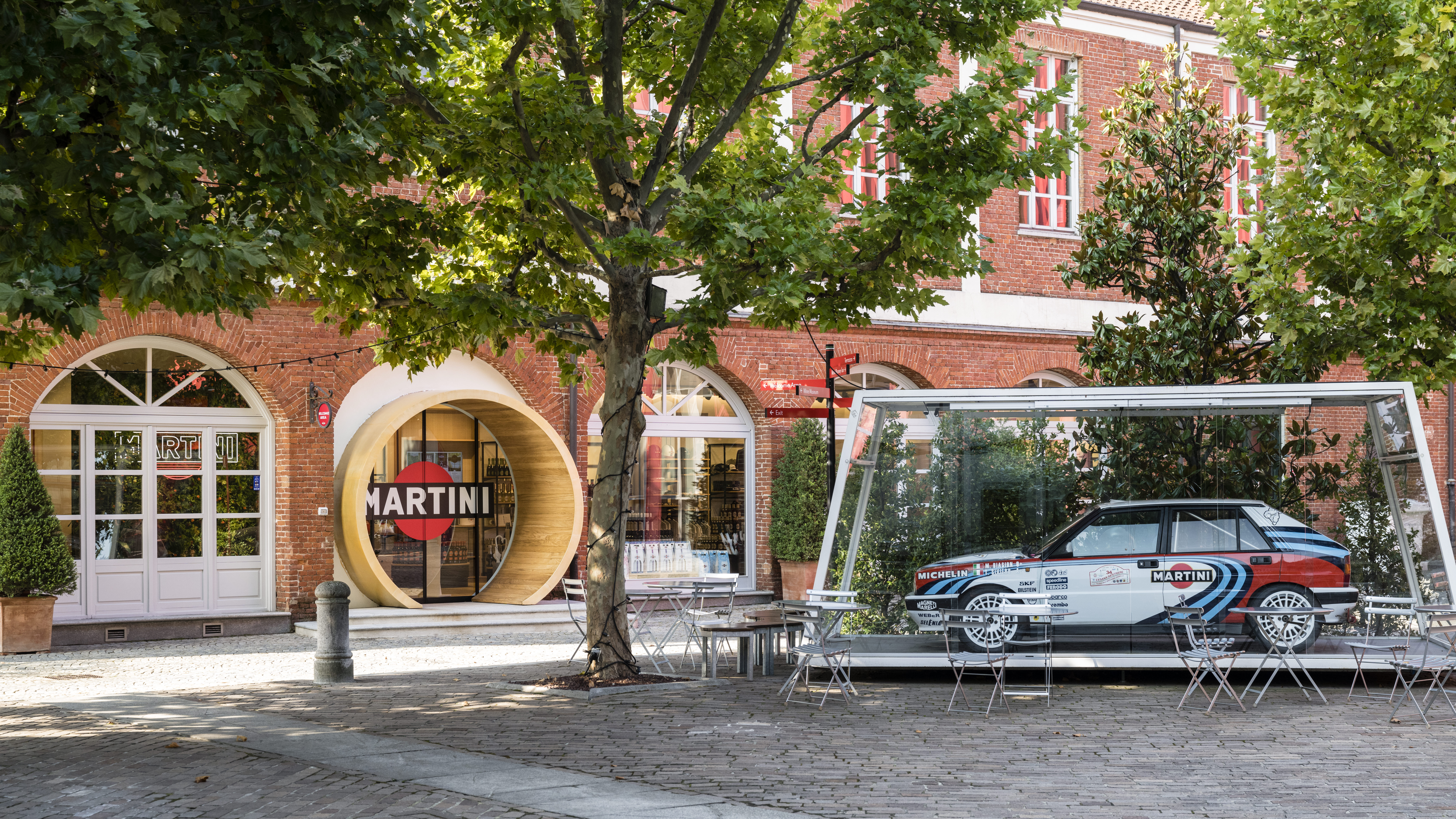 Drinking, cocktails, Italy - Photo of a white Martini branded car parked outside a red-brick building lined with trees.