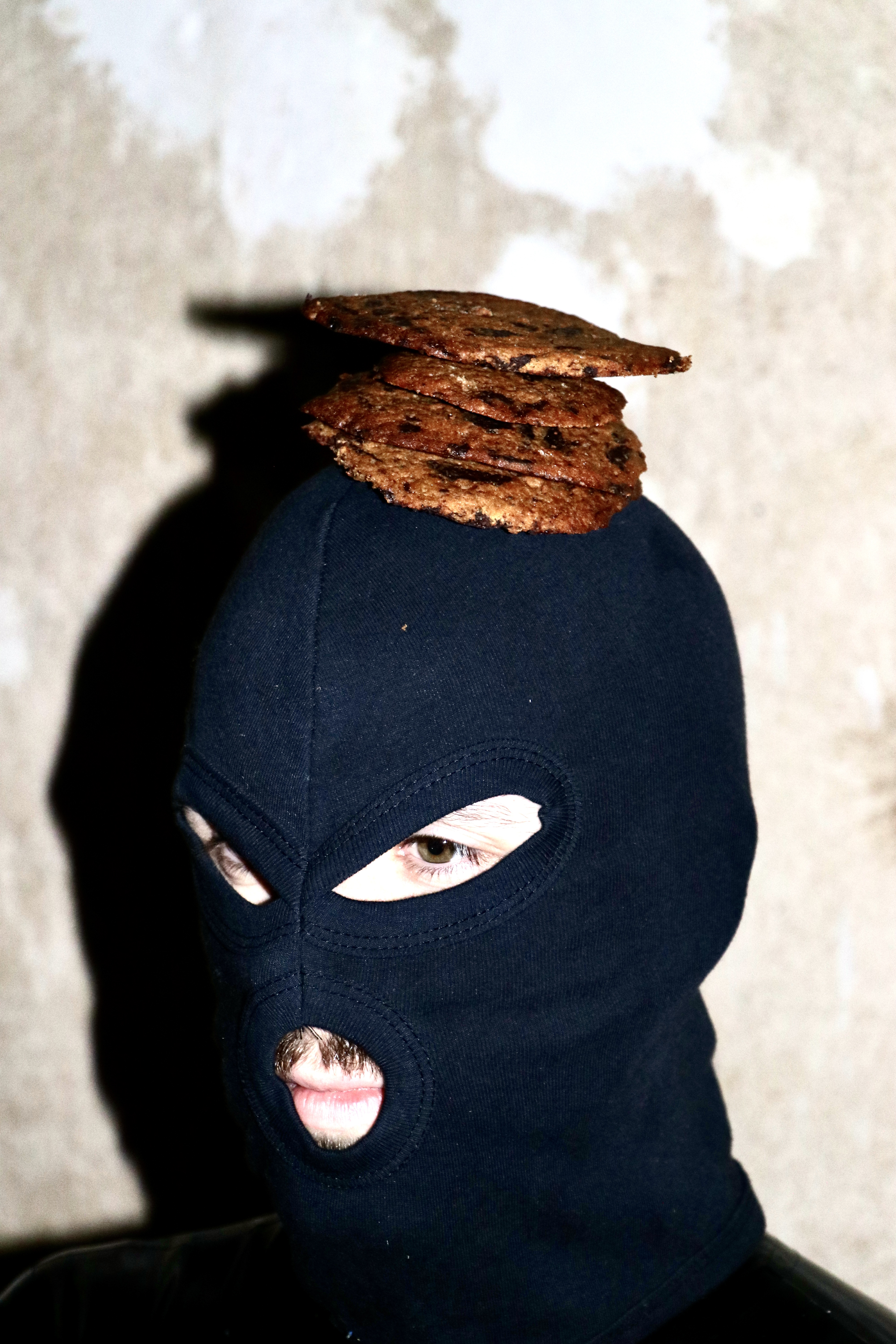 Kink, food, photography - Photograph of a man in a balaclava with four thin, brown biscuits resting on top of his head.