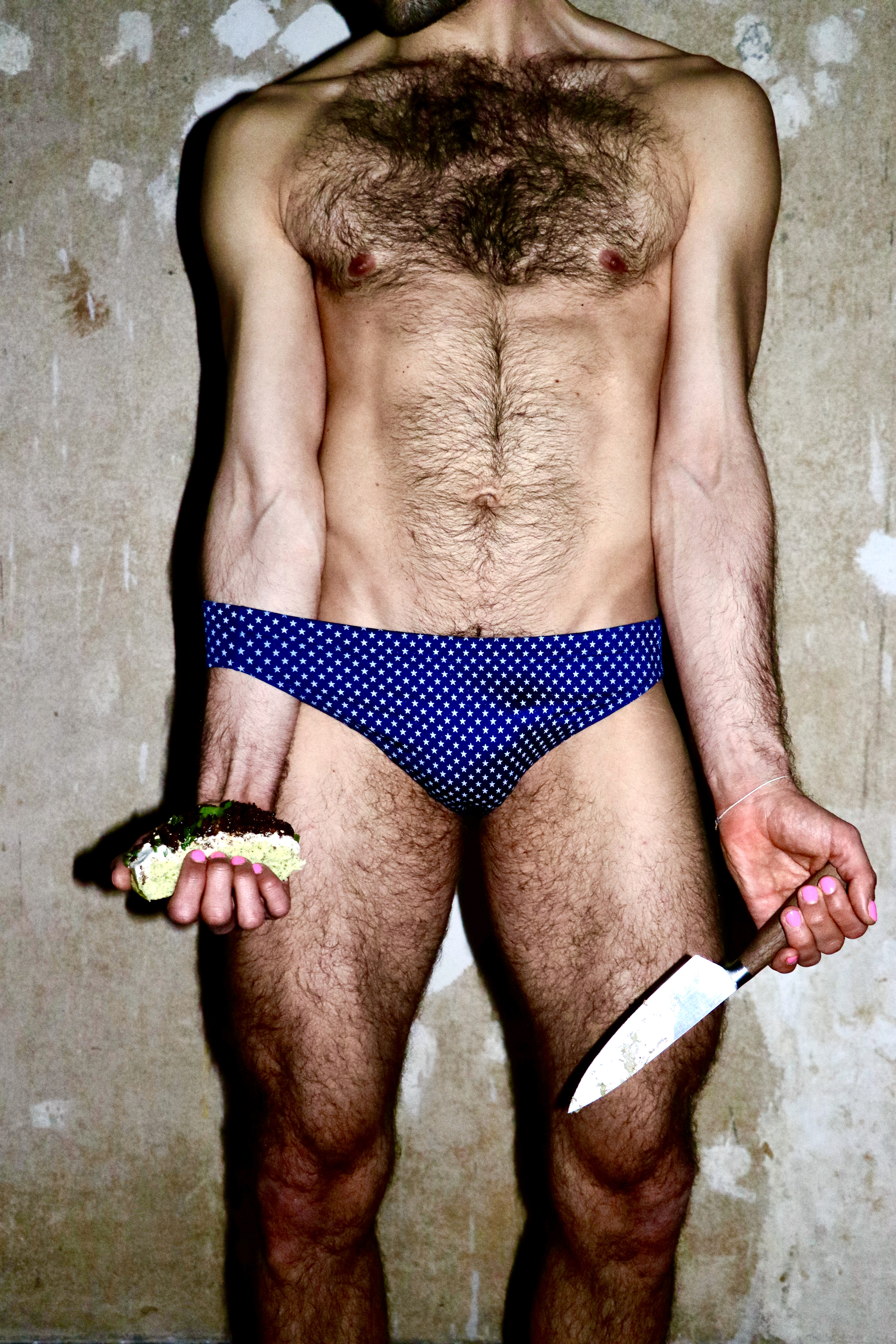 Kink, food, photography - Photograph of a hair man in a pair of blue swimming trunks holding a knife in one hand and a doughnut in the other.