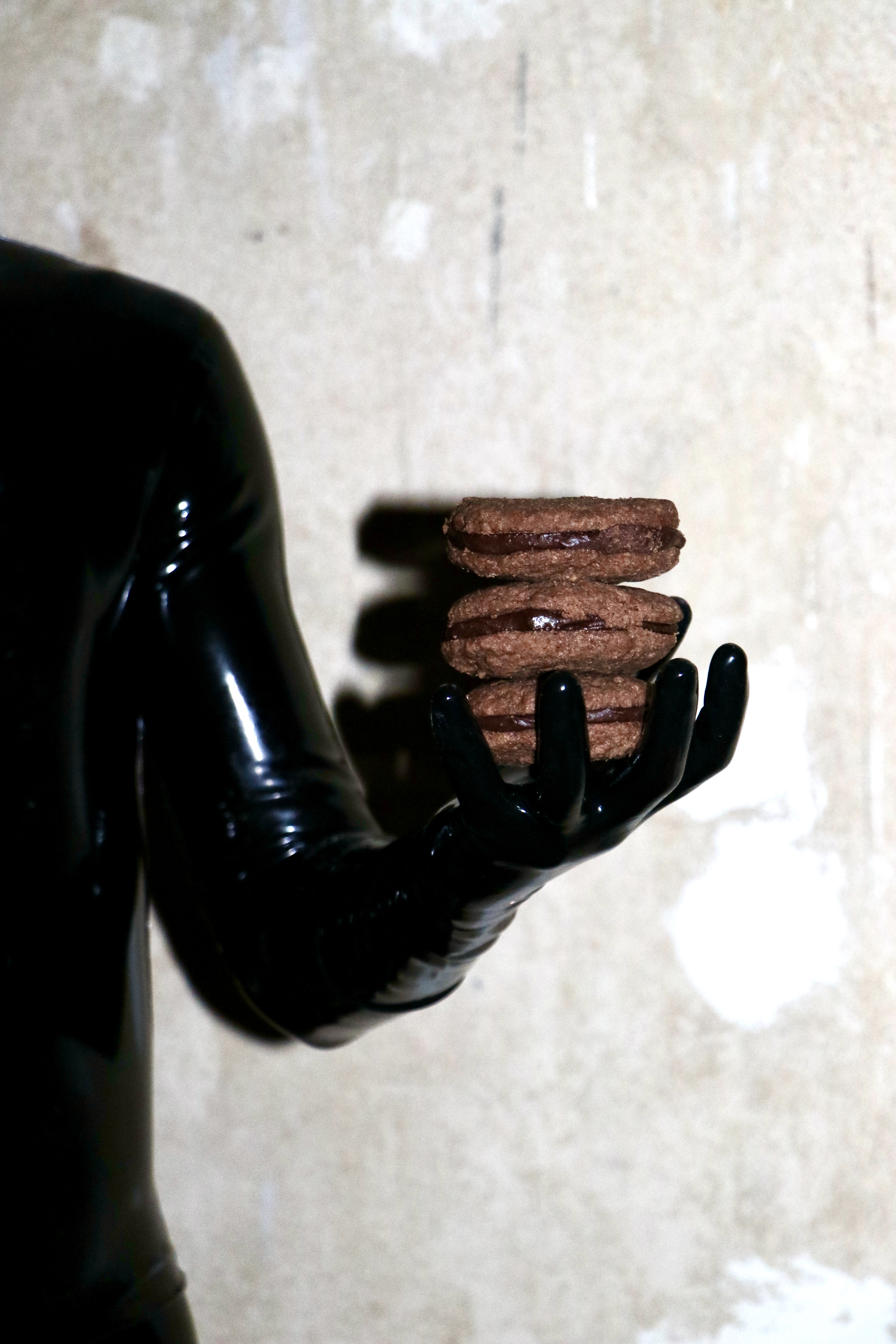 Kink, food, photography - Photograph of headless human figure wearing a black latex bodysuit holding a pile of three chocolate cookies.