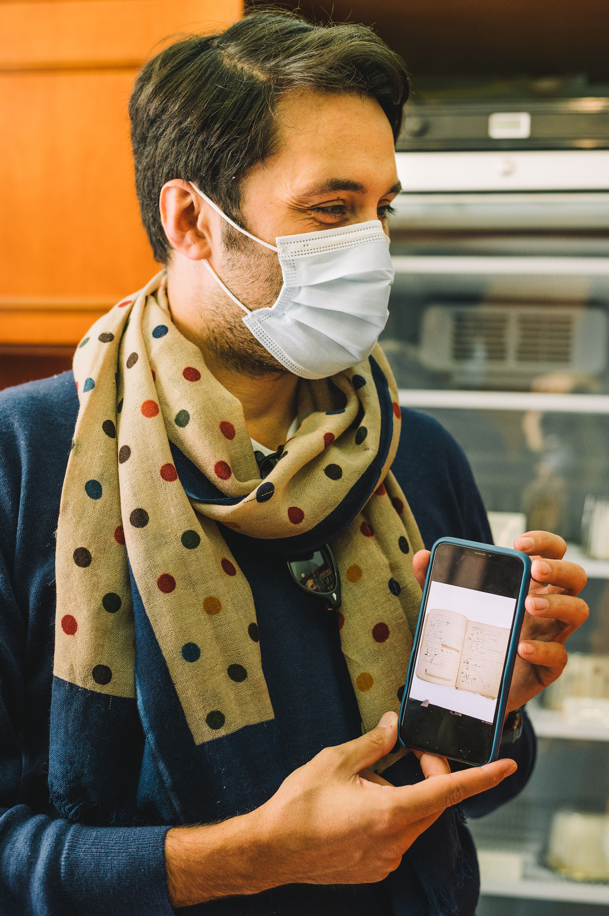 Mokambo, ice cream, Italy - Photograph of a man in a blue sweater draped with a cream and dotted scarf, holding up a mobile phone showing a scan of an old, lined notebook.