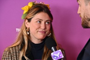 Photo shows Caroline Calloway with flowers in her hair against a pink backdrop. She's being interviewed by man holding a microphone who's half out of frame. Her lips are pursed.