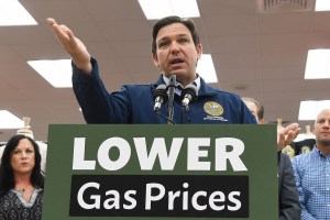 Florida Gov. Ron DeSantis stands in front of a sign that says LOWER GAS PRICES