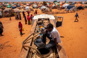 A truck driver sits on top of the water truck as it pumps water to a water point