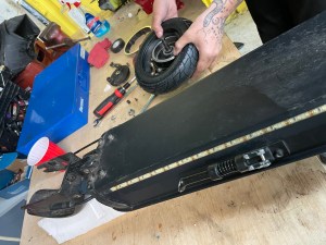 An electric scooter sits on a wooden work bench with its back wheel removed. A pair of hands pulls a busted tire off from the rim.