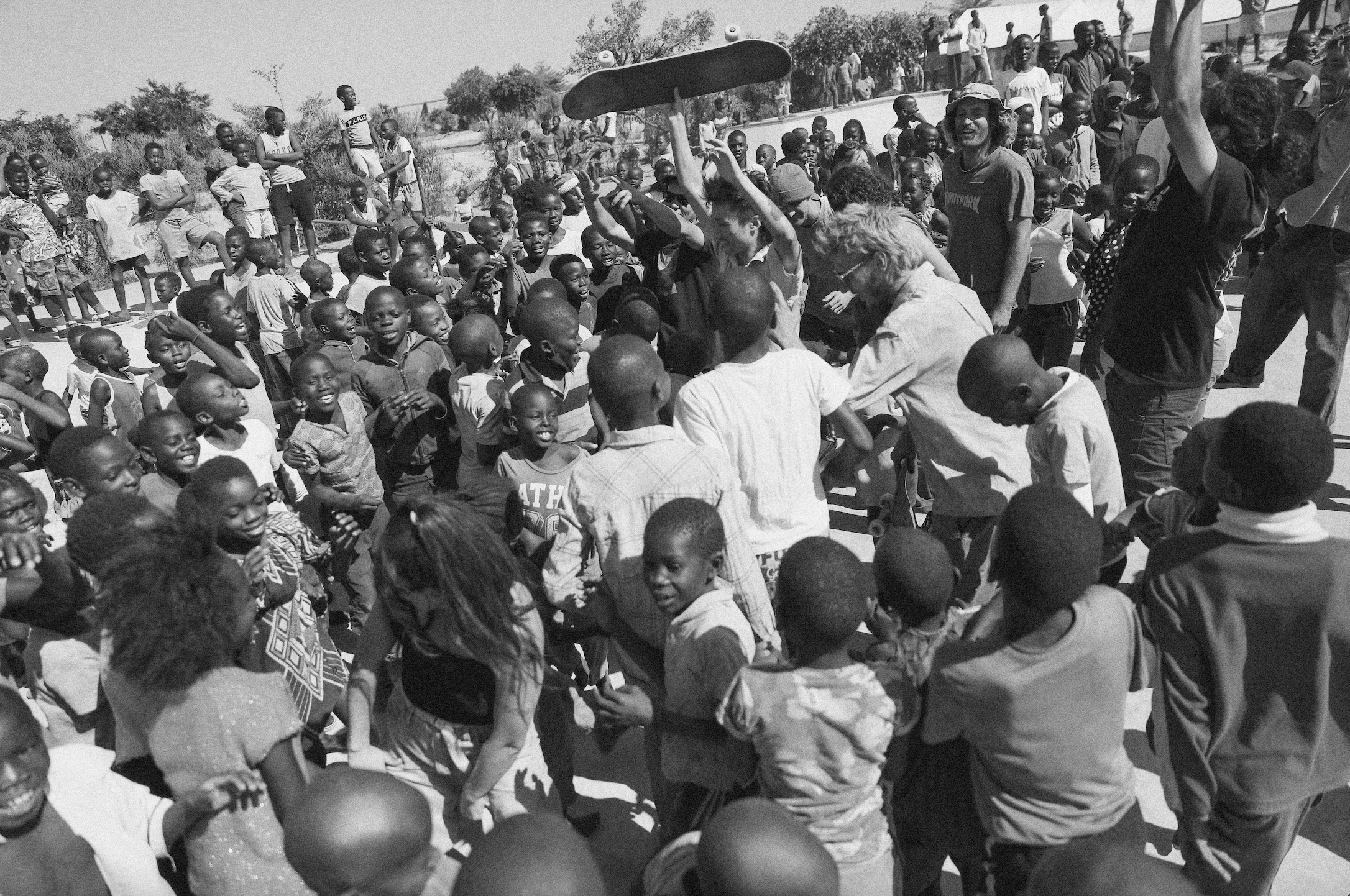 Africa, skateboarding, Jonas Camps - Black and white photo of a large group of youths looking happily at a man holding a skateboard above his head.