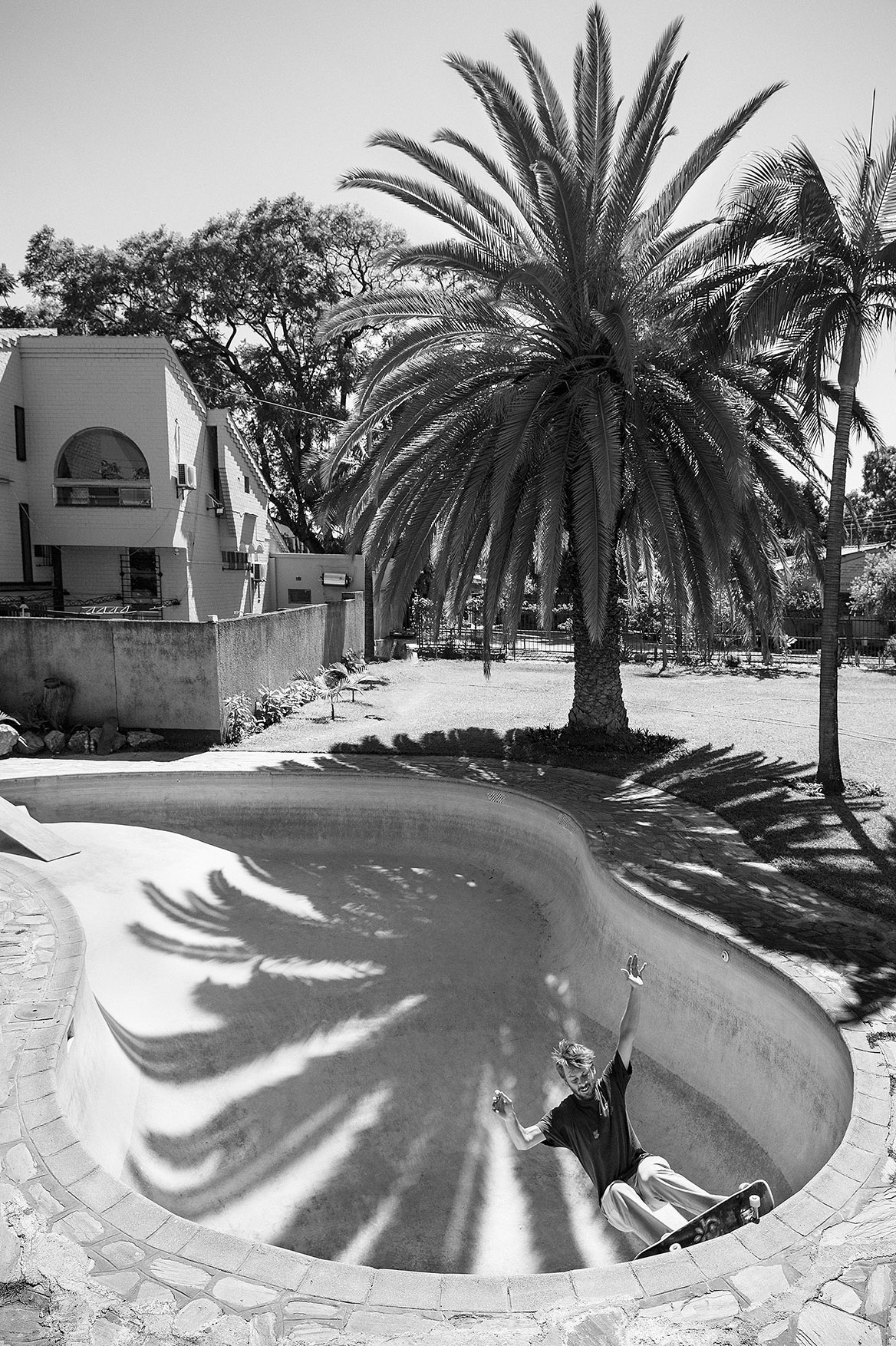 Africa, skateboarding, Jonas Camps - Black and white photo of a man skating in an empty swimming pool, fringed by palm trees.