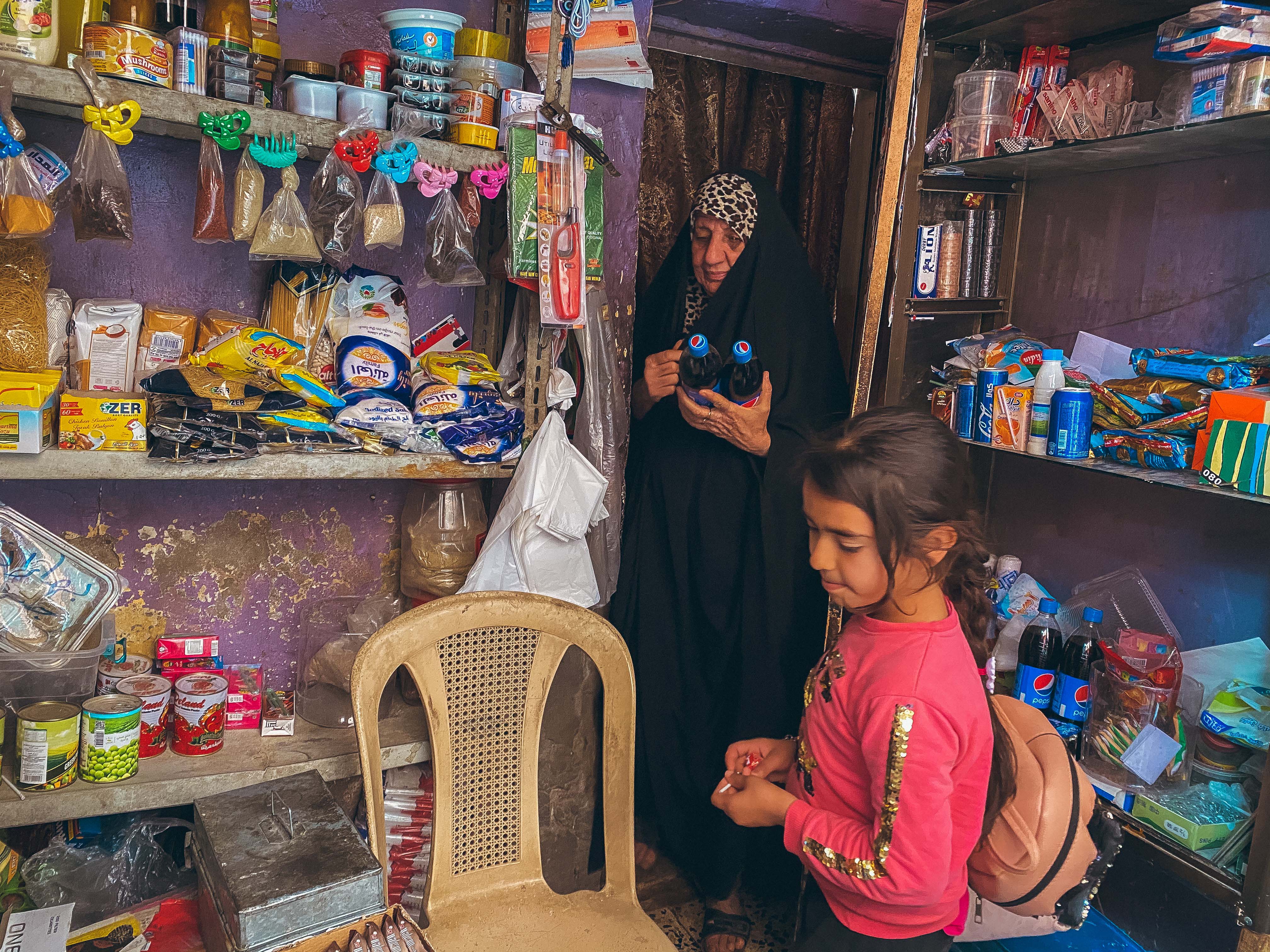 Kadhimiya district, Baghdad – Middle-aged woman wearing a long black abaya and a leopard headscarf inside a small shop filled with food and other household products. She's about to give two bottles of pepsi to a little girl wearing a pink and gold top.