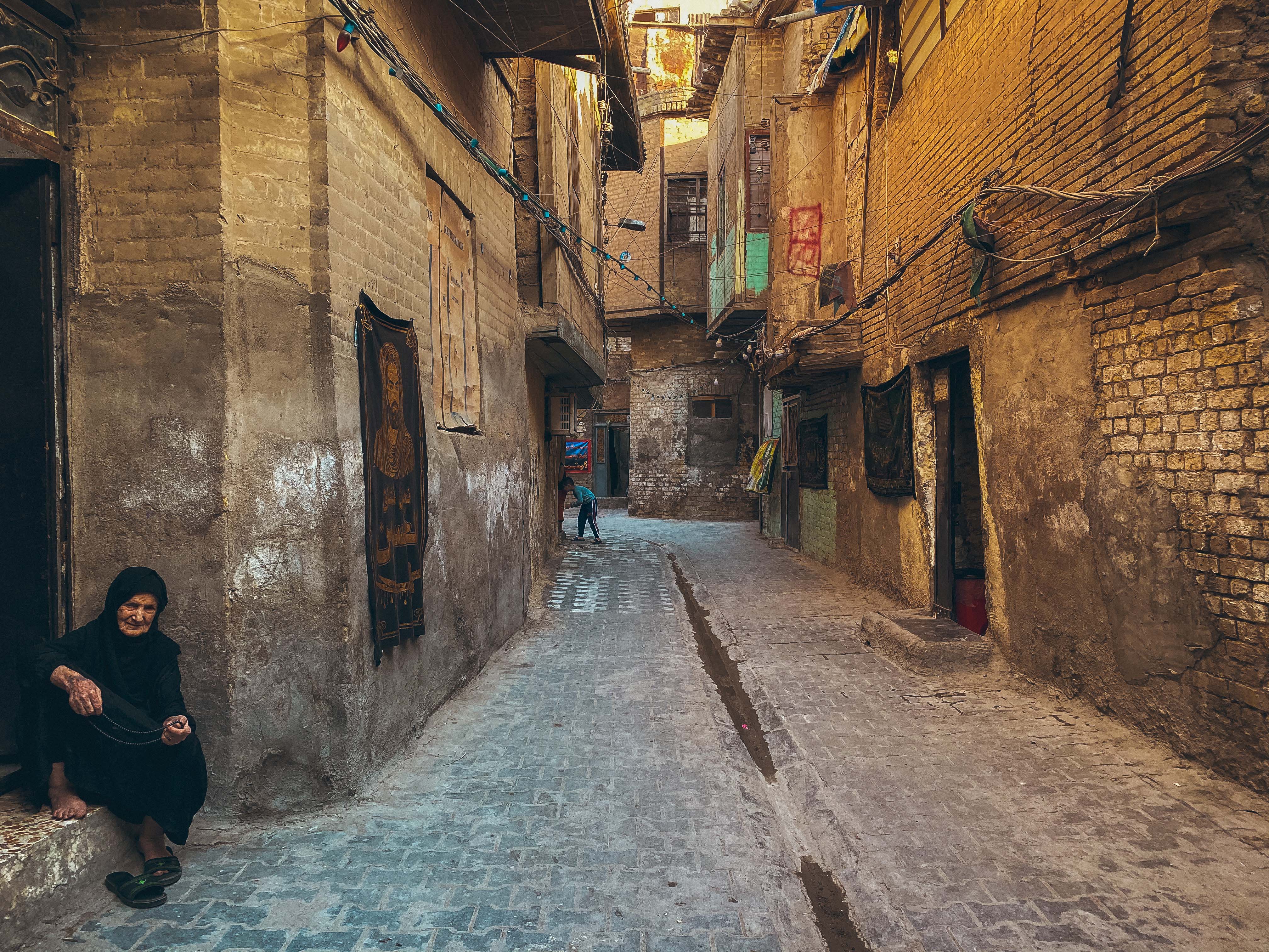 Sadriya District, Baghdad – a narrow, cobble-stone street. In the foreground, an old lady is sitting on the step at the entrance of her house.