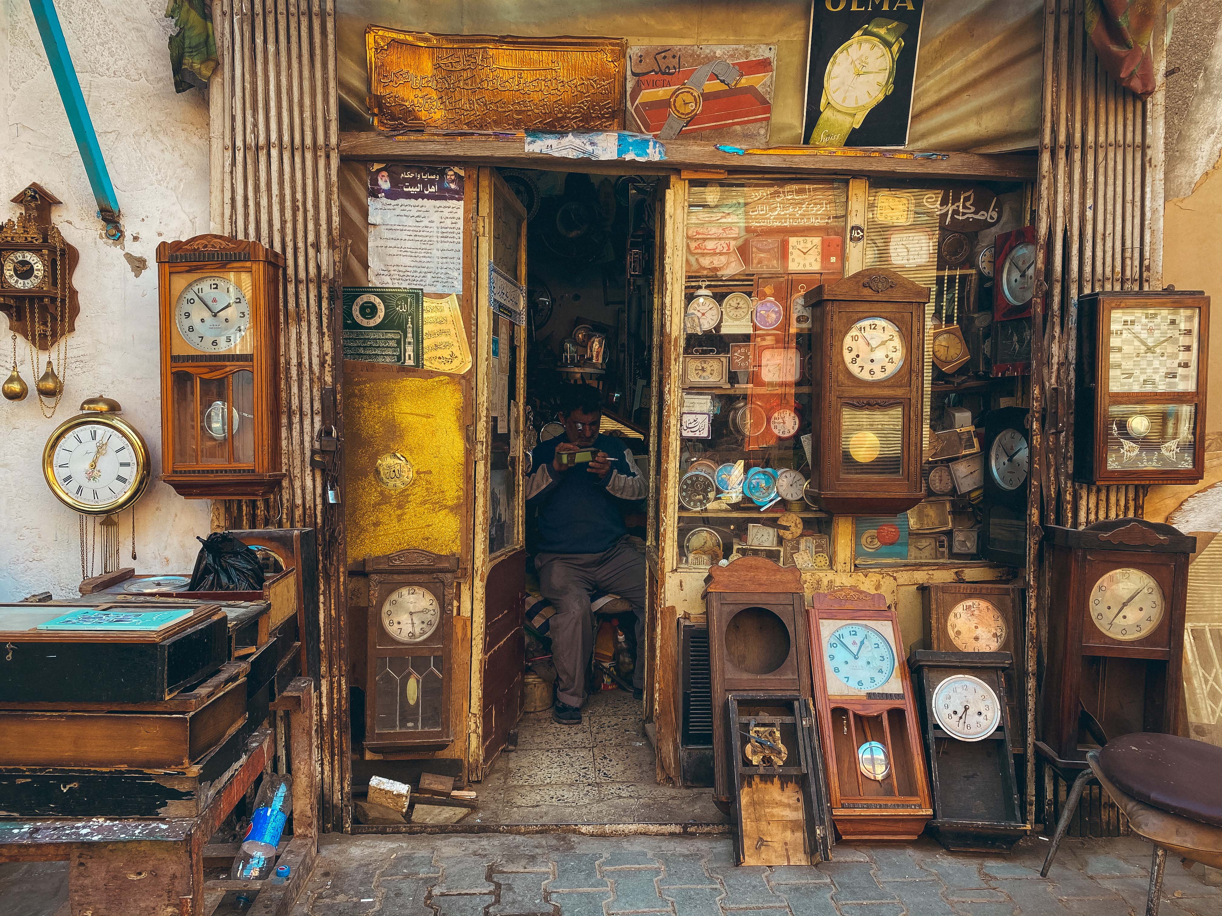 Al-Midan District, Baghdad – view from outside of narrow shop filled with clocks. Inside, a man is looking at a watch with a magnifying monocle.