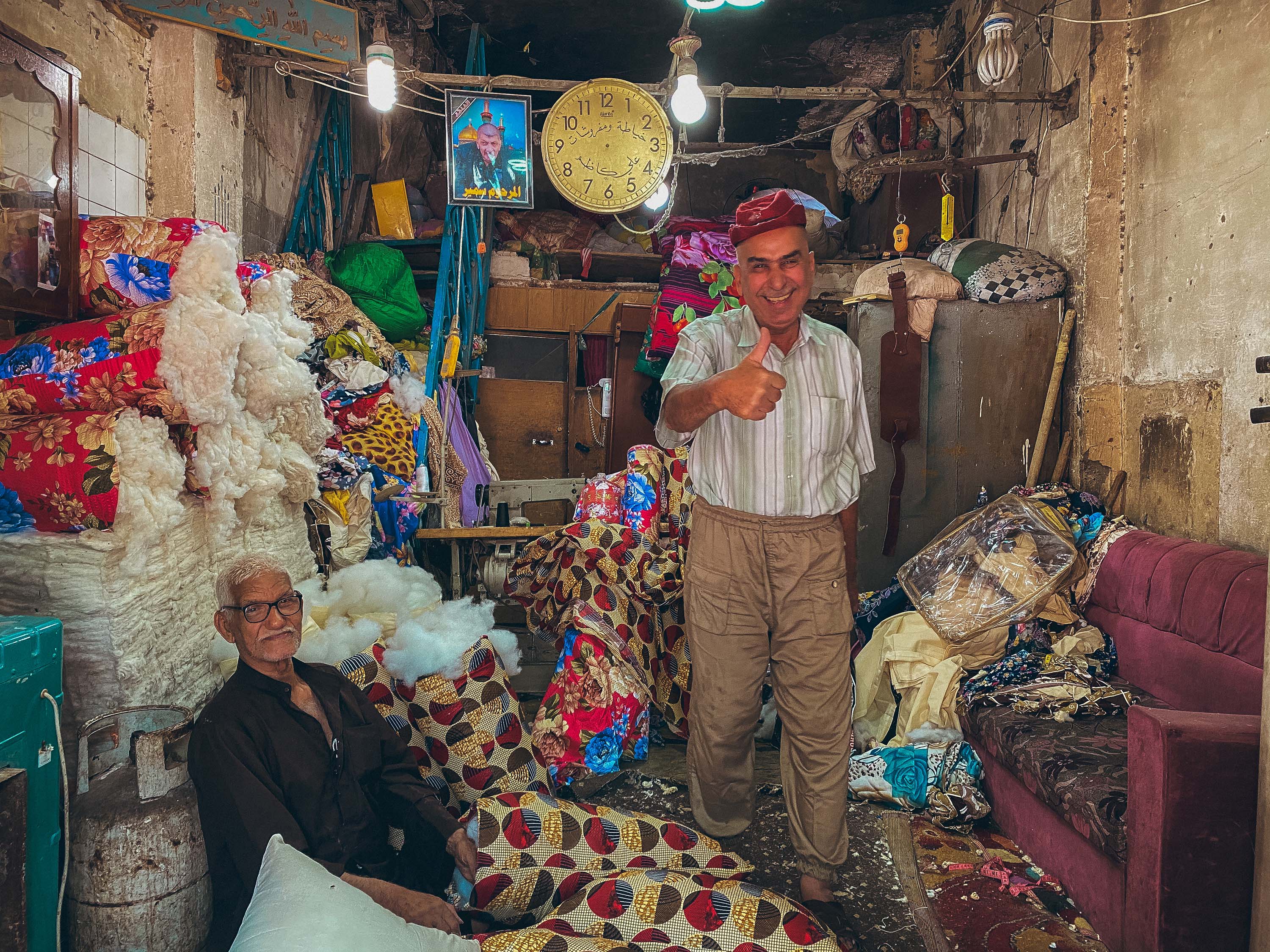 Sadriya Market, Baghdad – two old men, one sitting the other standing in the middle of a shop filled with old furniture and couch stuffing. One of them is smiling and giving the photographer the thumbs up.