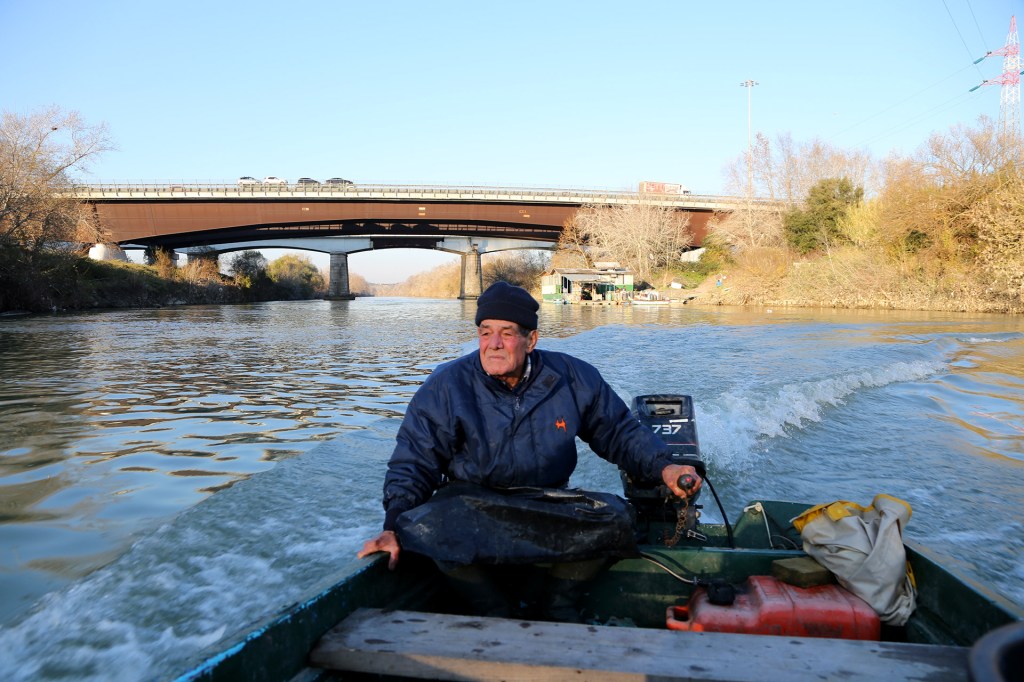 Sconocchia, Tiber, Rome – Man driving a small boat on the river, wearing a blue wooly hat and a winter jacket and looking at the distance.