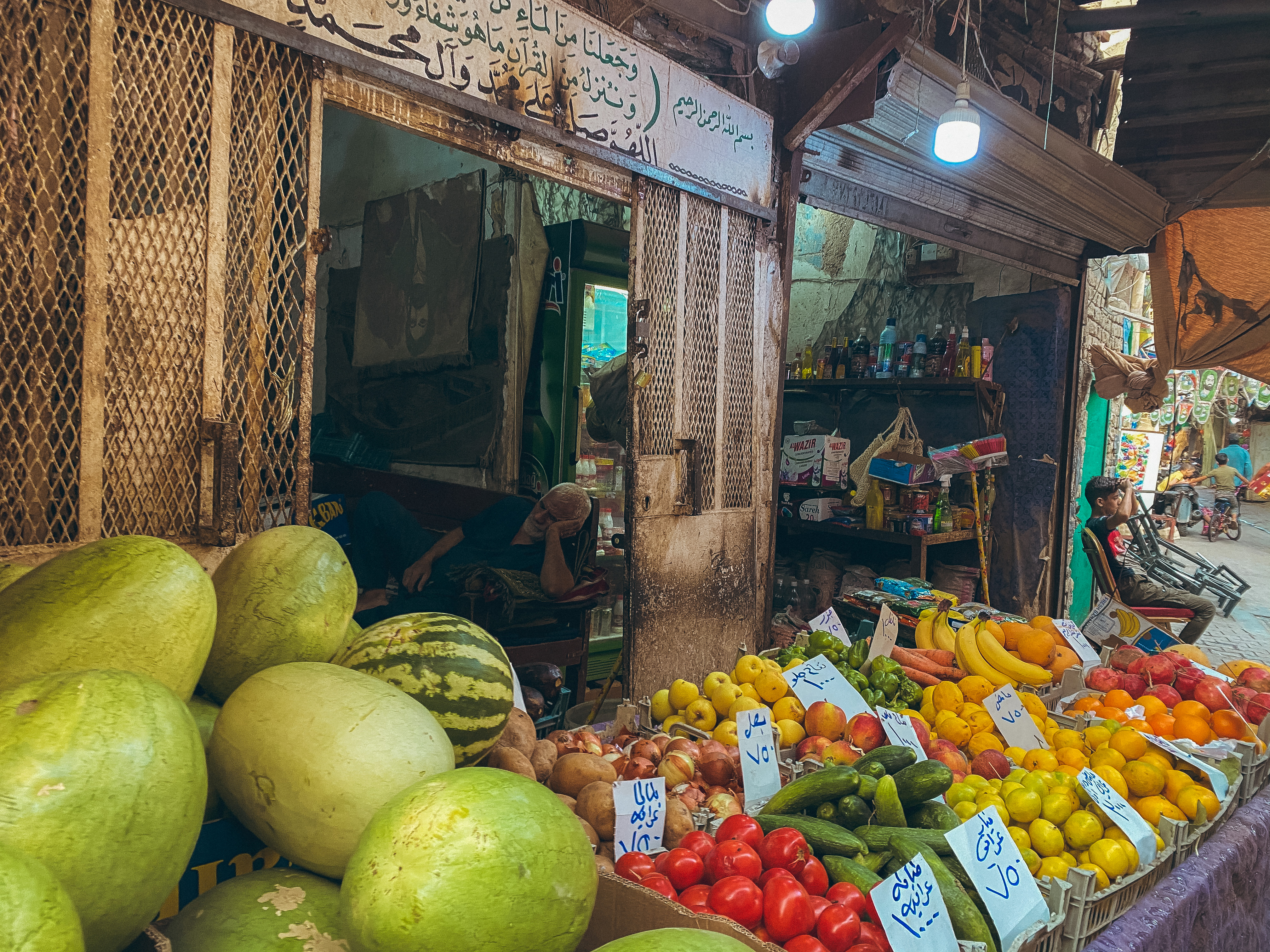 Sadriya Market, Baghdad – foreground: fruits and vegetables, including cucumber, tomatoes and watermelon. Background: old man napping on a couch.