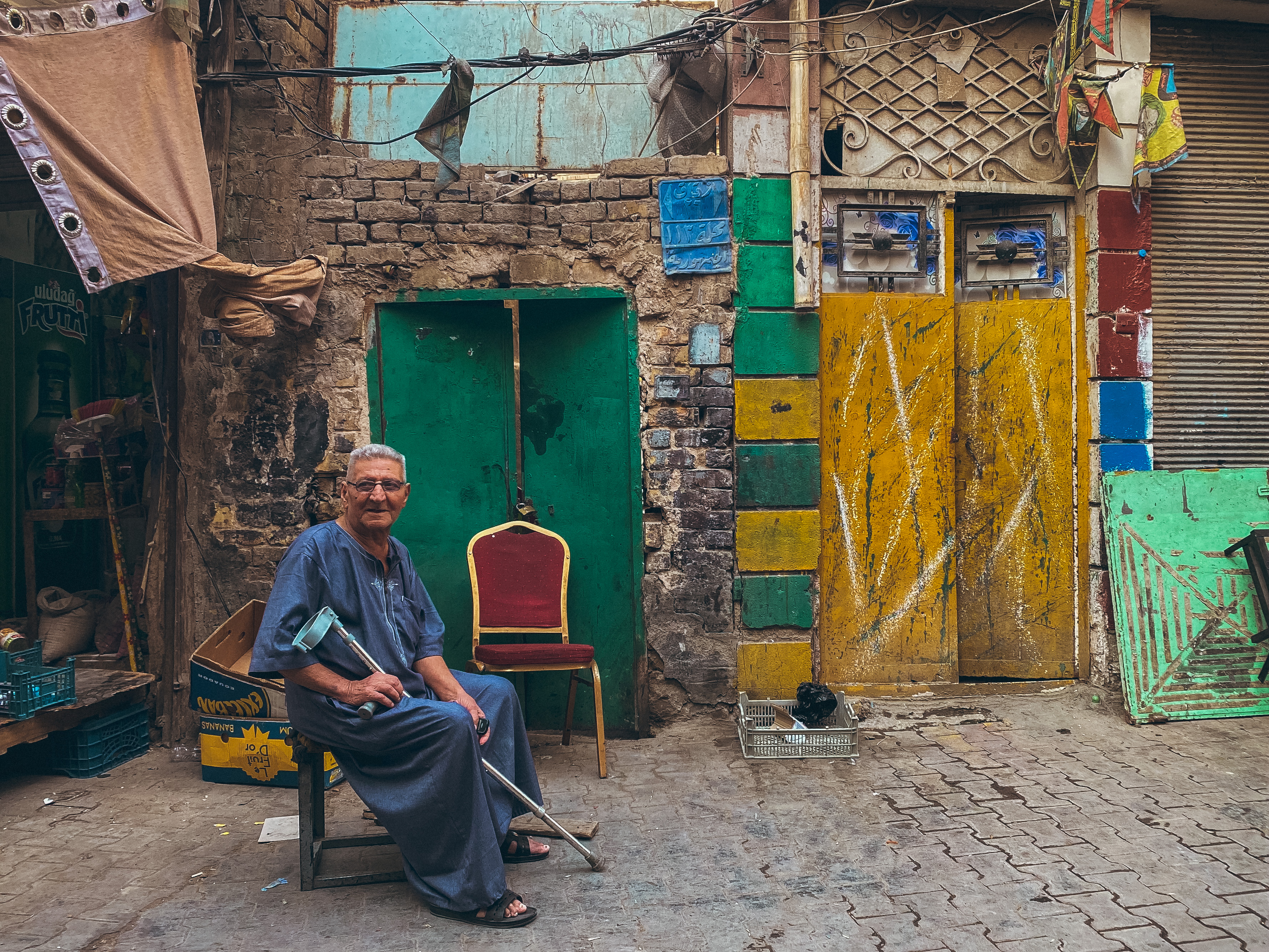 Sadriya district, Baghdad – old man wearing a long blue robe, sitting on a chair in the street with one crutch.