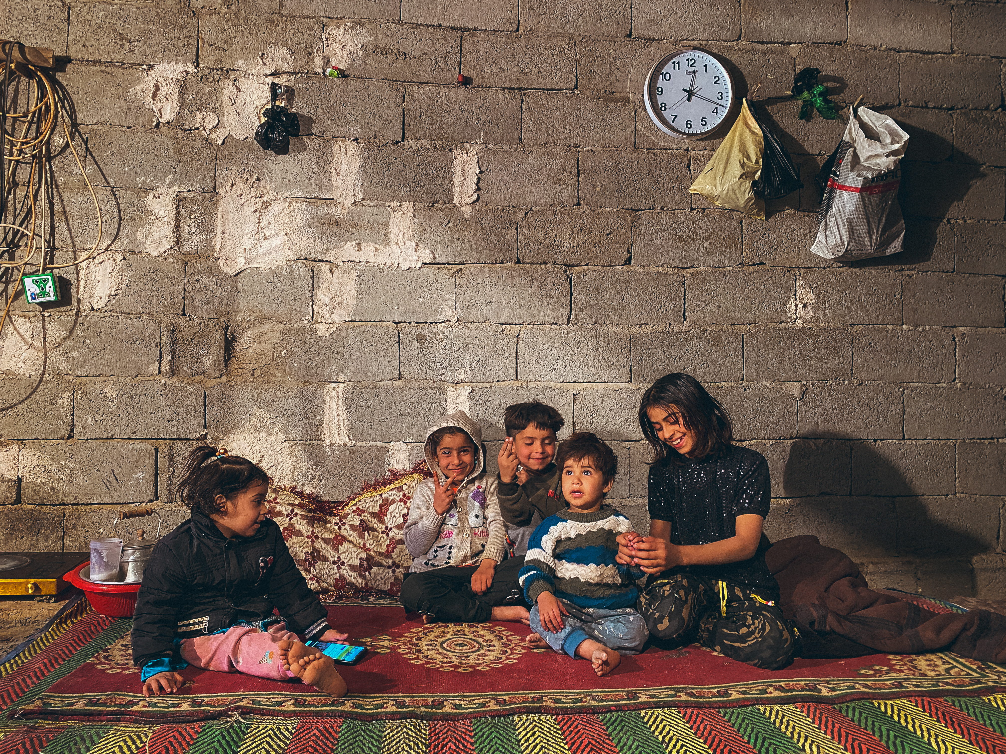 Nahrawan district, Baghdad – three young boys, a young girl and a teenager sitting on a large red carpet in front of a grey wall and interacting with each other.