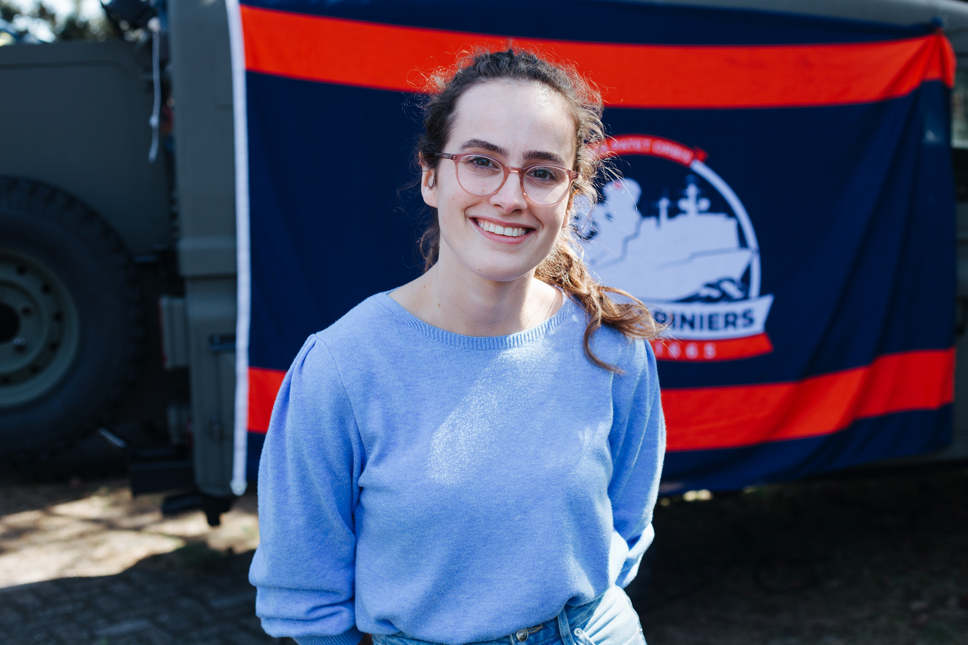 Army recruitment, Fort 1881 – young woman with long brown hair and glasses, posing in front of a red and blue flag.