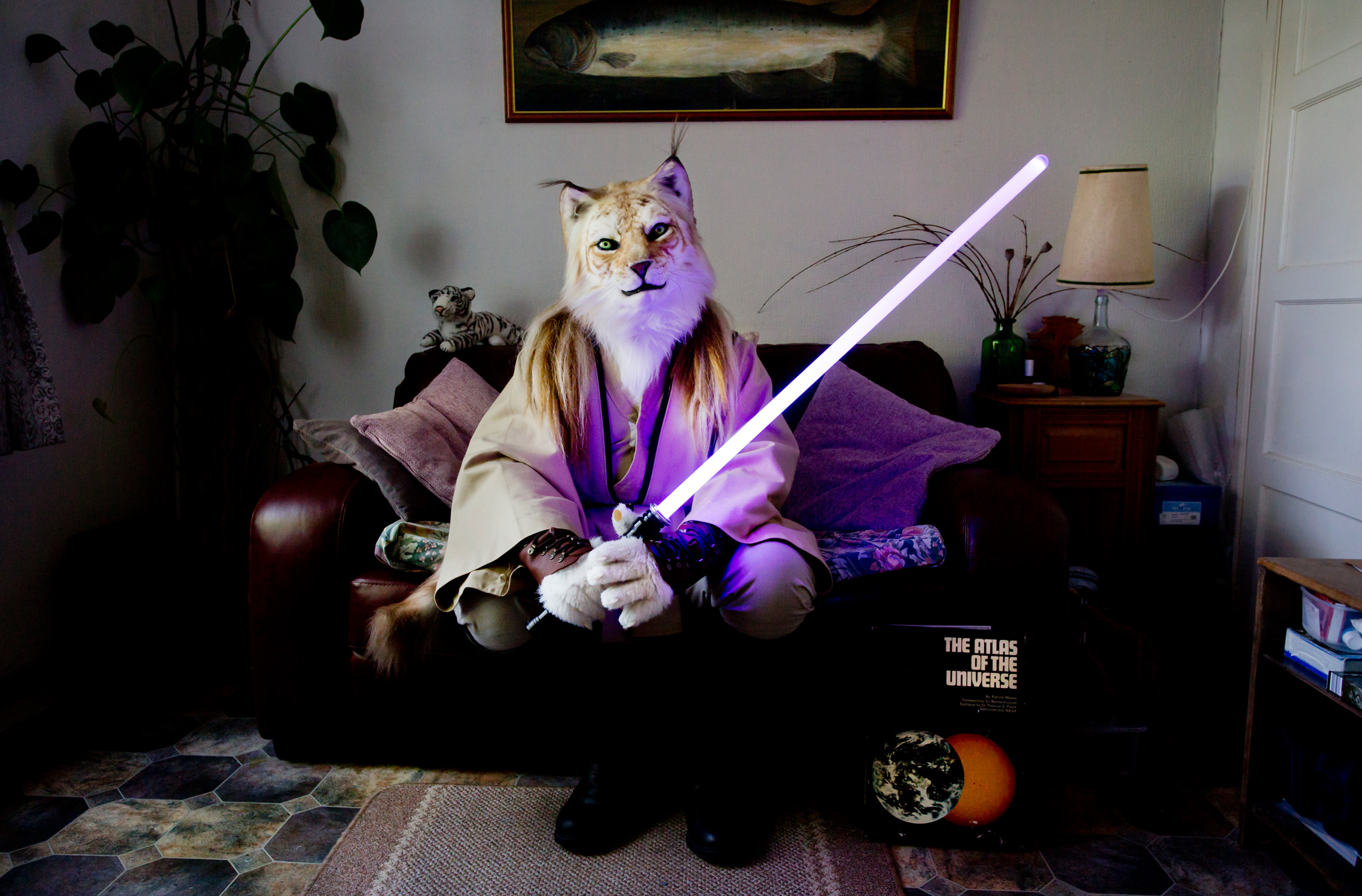 A fursuiter dressed as a Jedi tiger sits at home on their sofa, holding a light saber.