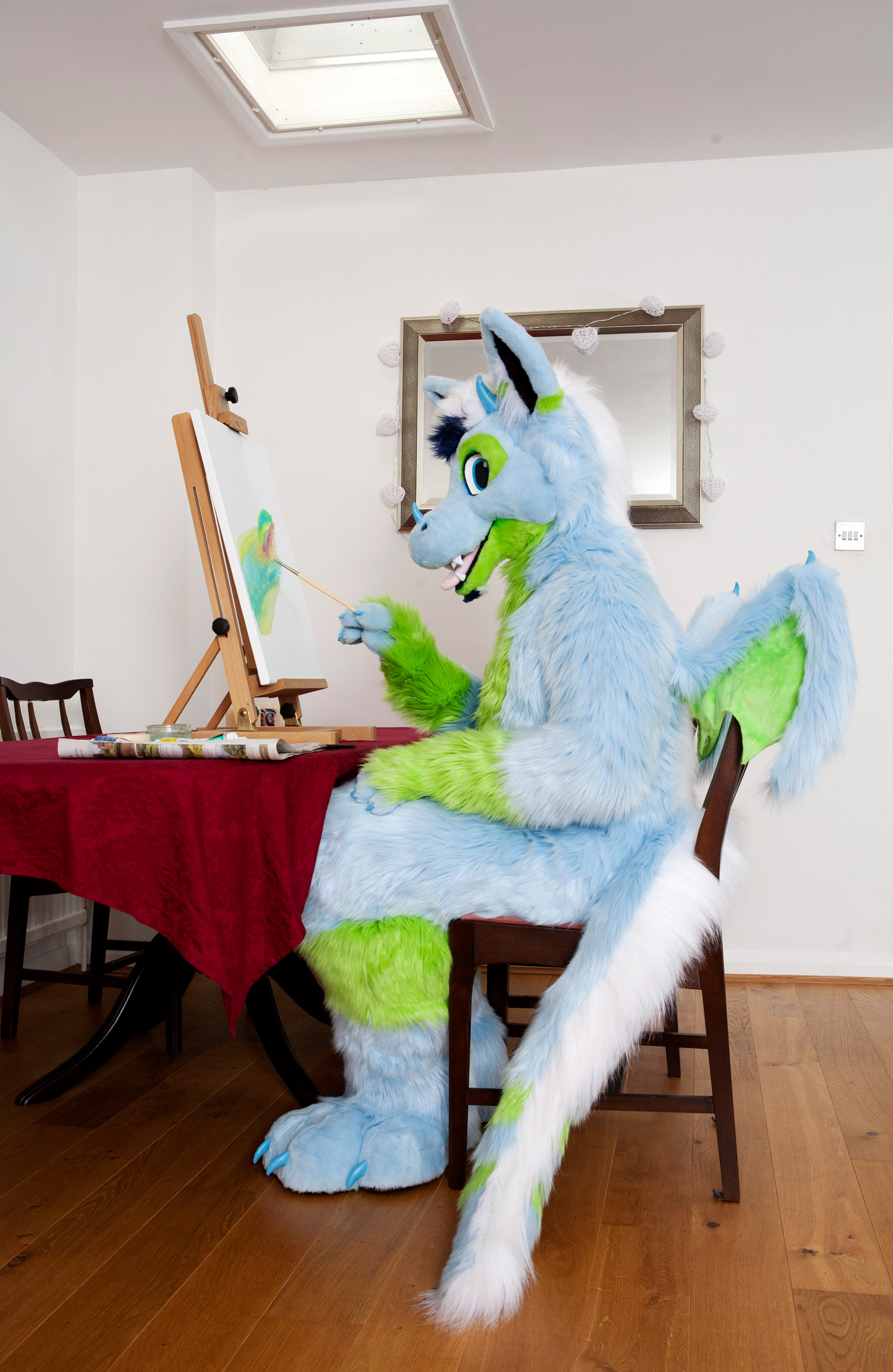 A fursuiter dressed as a dragon sits down at the table to do some painting.