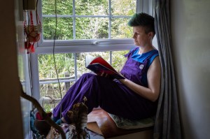 A queer gender non-conforming woman reading a book by a window in